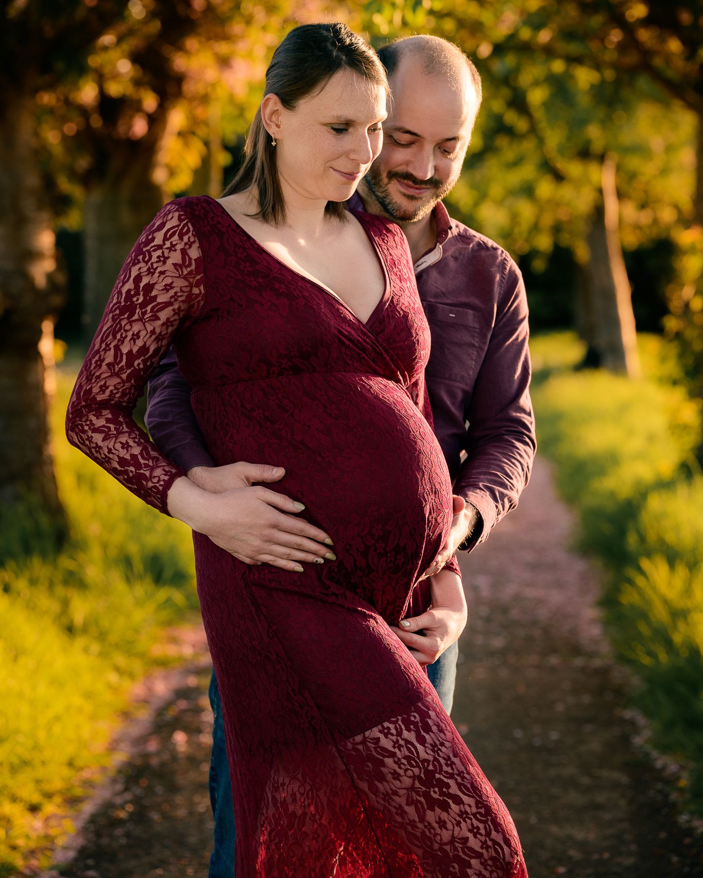Couple tenderly embracing pregnant woman in red lace maternity gown on sunlit garden path.