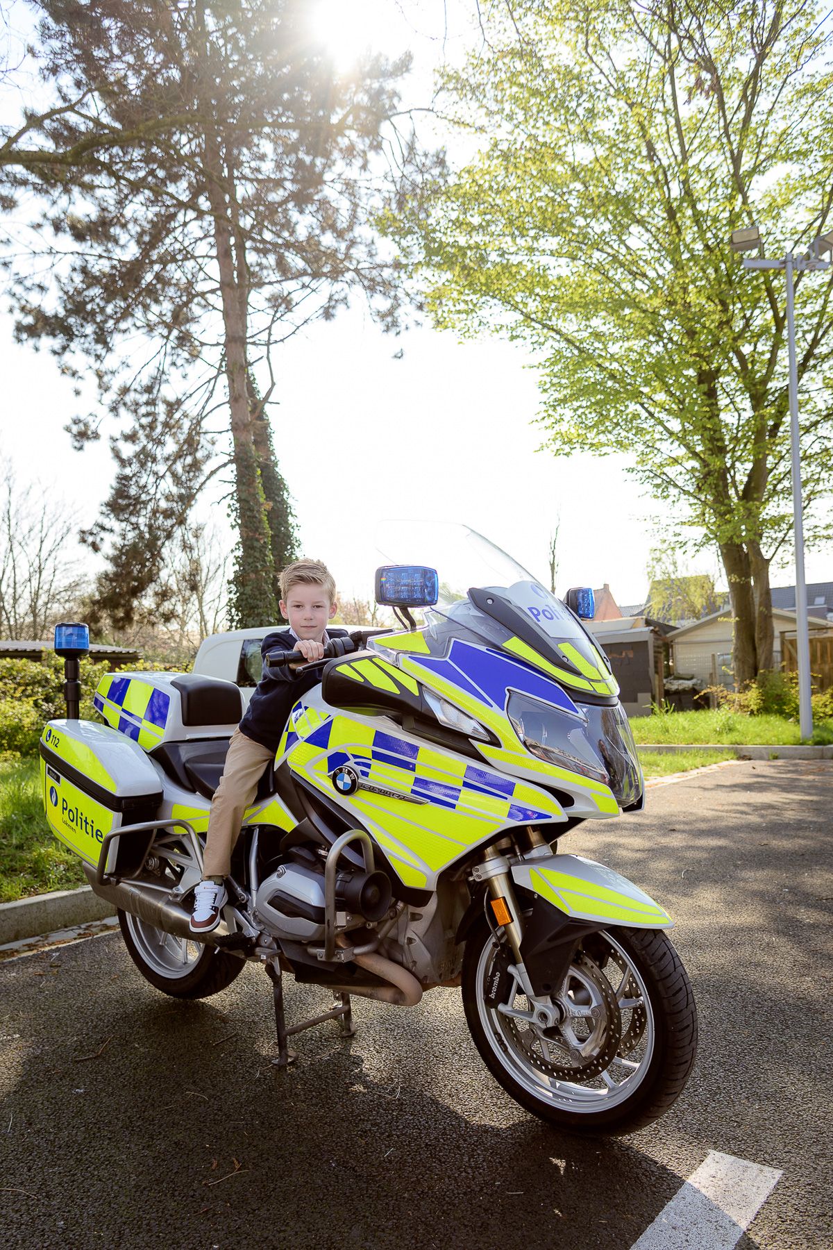 Young boy sitting on a yellow and green police motorcycle parked on a road surrounded by trees.