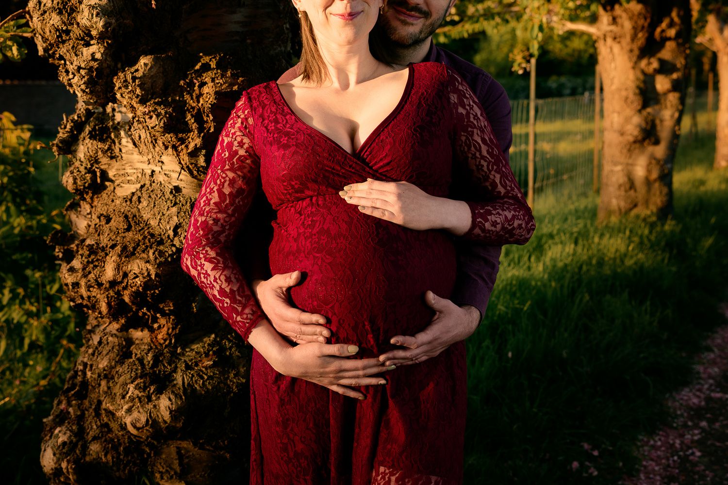Pregnant woman in red lace dress cradling baby bump with partner embracing her outdoors in golden sunlight.