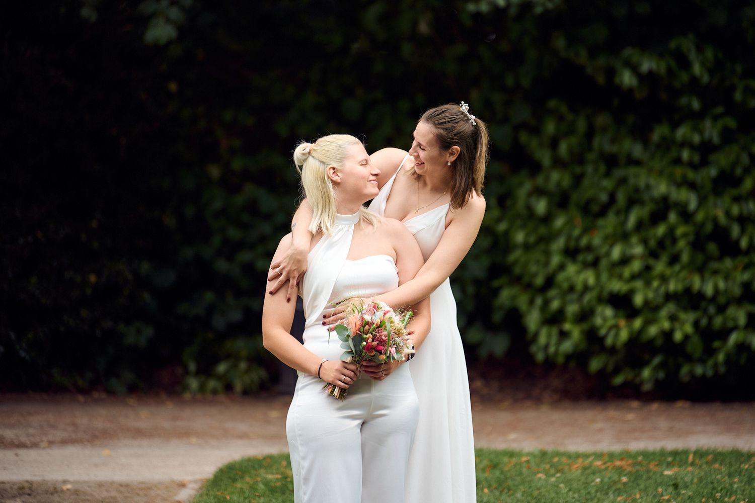 Two brides in white embracing outdoors, one holding a colorful bouquet, surrounded by lush green hedges.