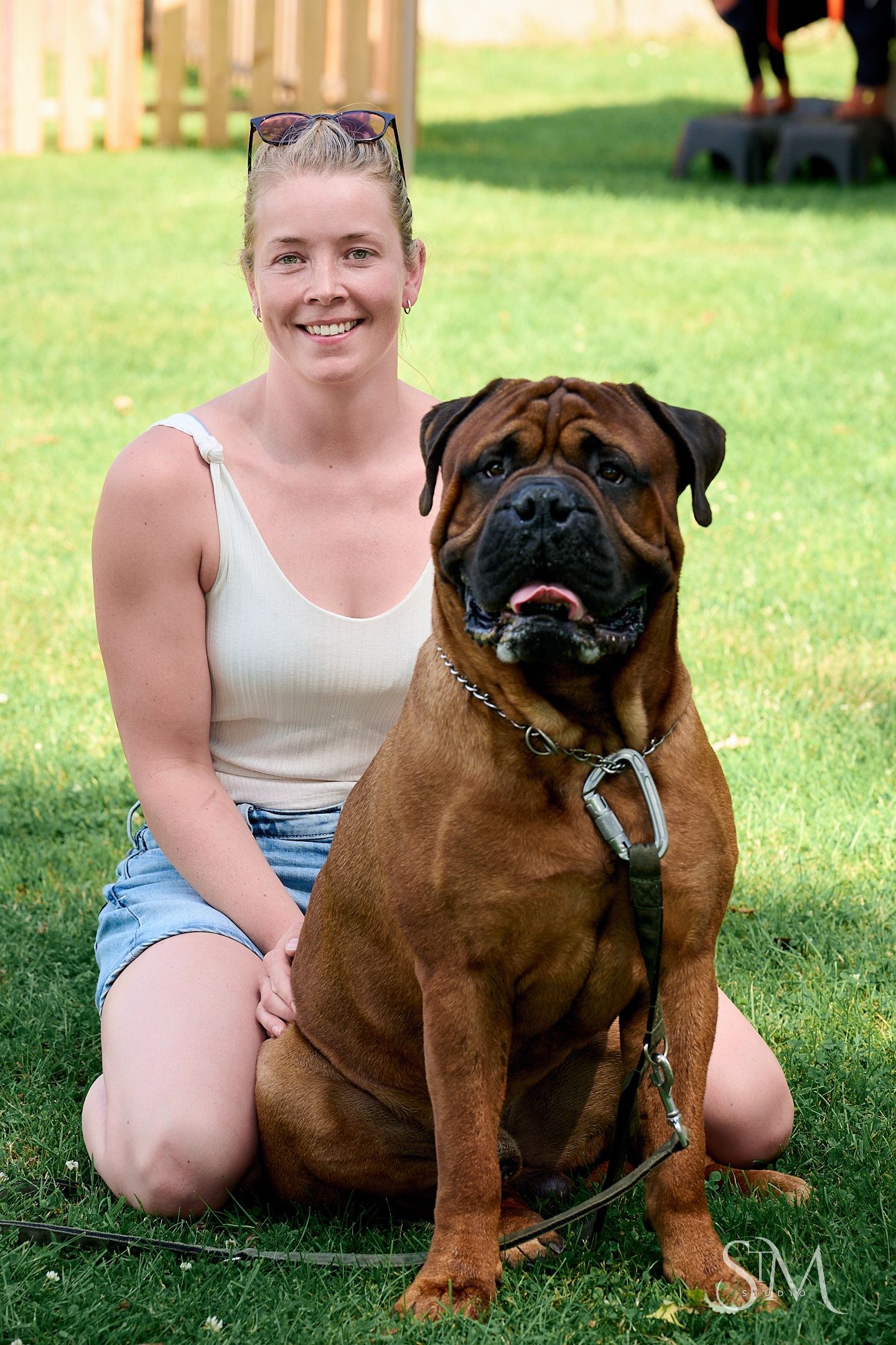 A smiling woman in a tank top sits on grass beside a large brown Bullmastiff dog on a leash outdoors.