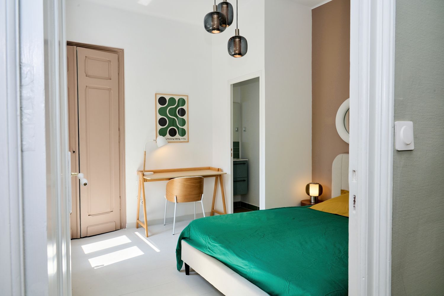 Modern bedroom with green bedspread, wooden desk, pendant lights, and minimalist decor in a bright white room.