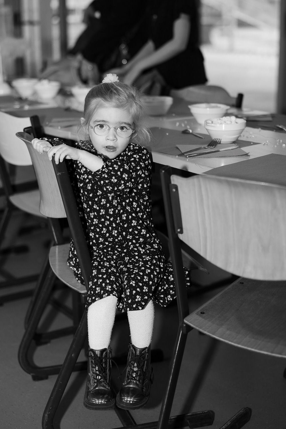 Young girl with glasses in a floral dress and boots sits alone at a dining table in black and white.