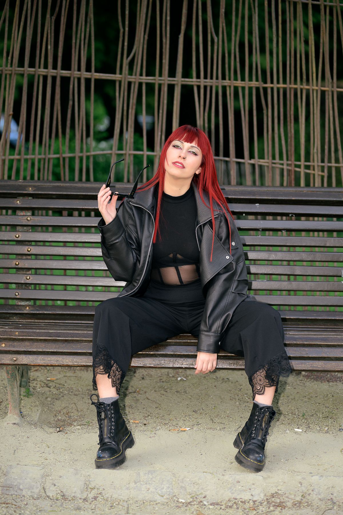 Red-haired woman in black leather jacket, lace pants, and boots sitting on a bench outdoors.