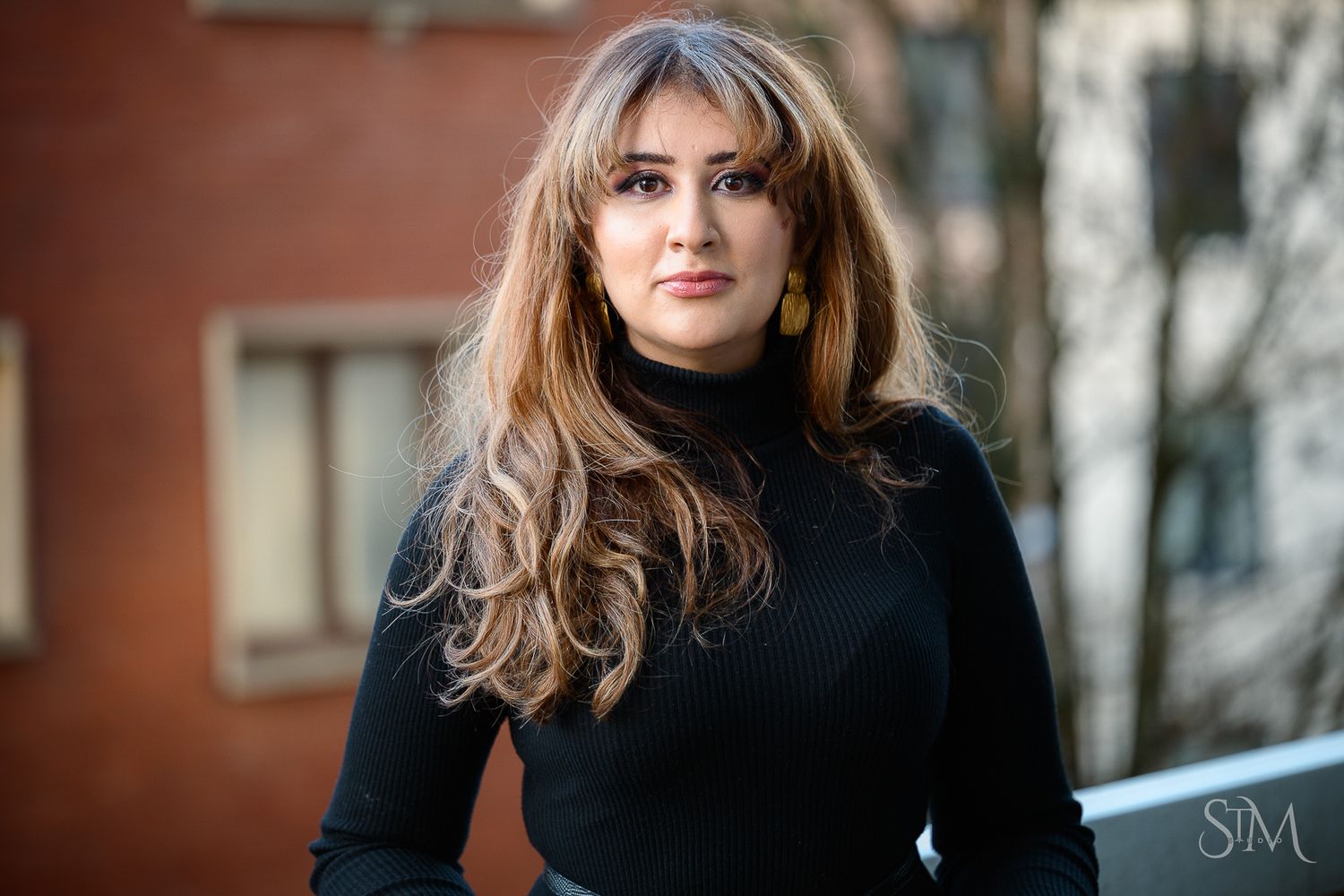 Young woman with long wavy hair wearing a black turtleneck sweater posing outdoors in an urban setting.