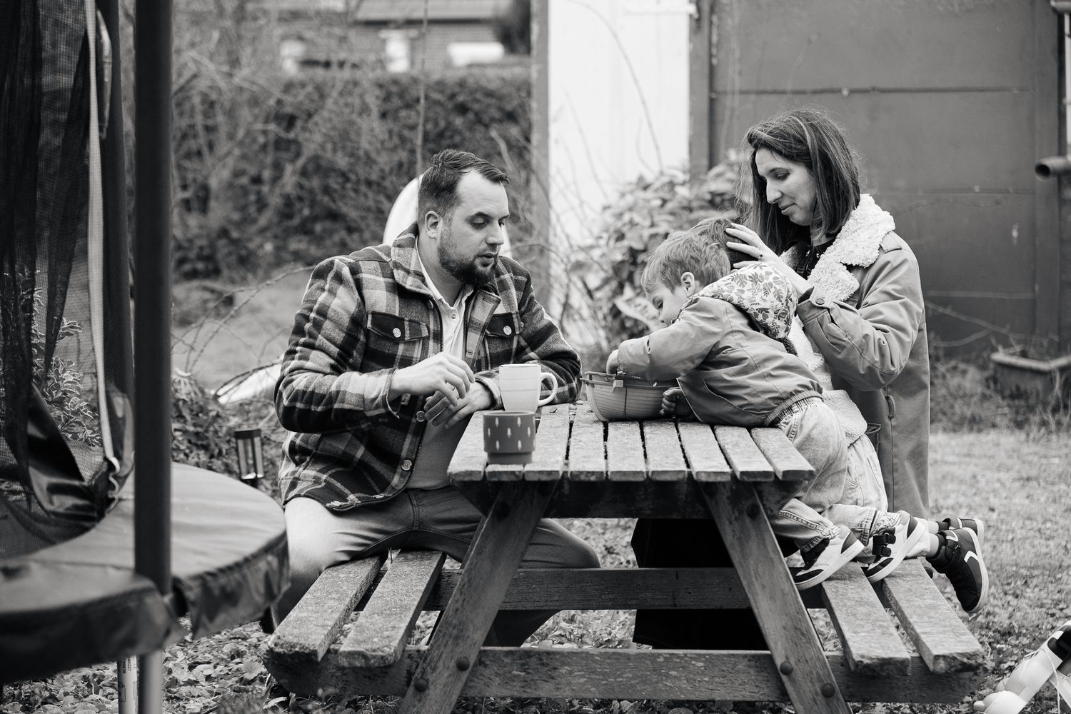 Black and white photo of a family with a young child sitting at an outdoor picnic table in a garden.