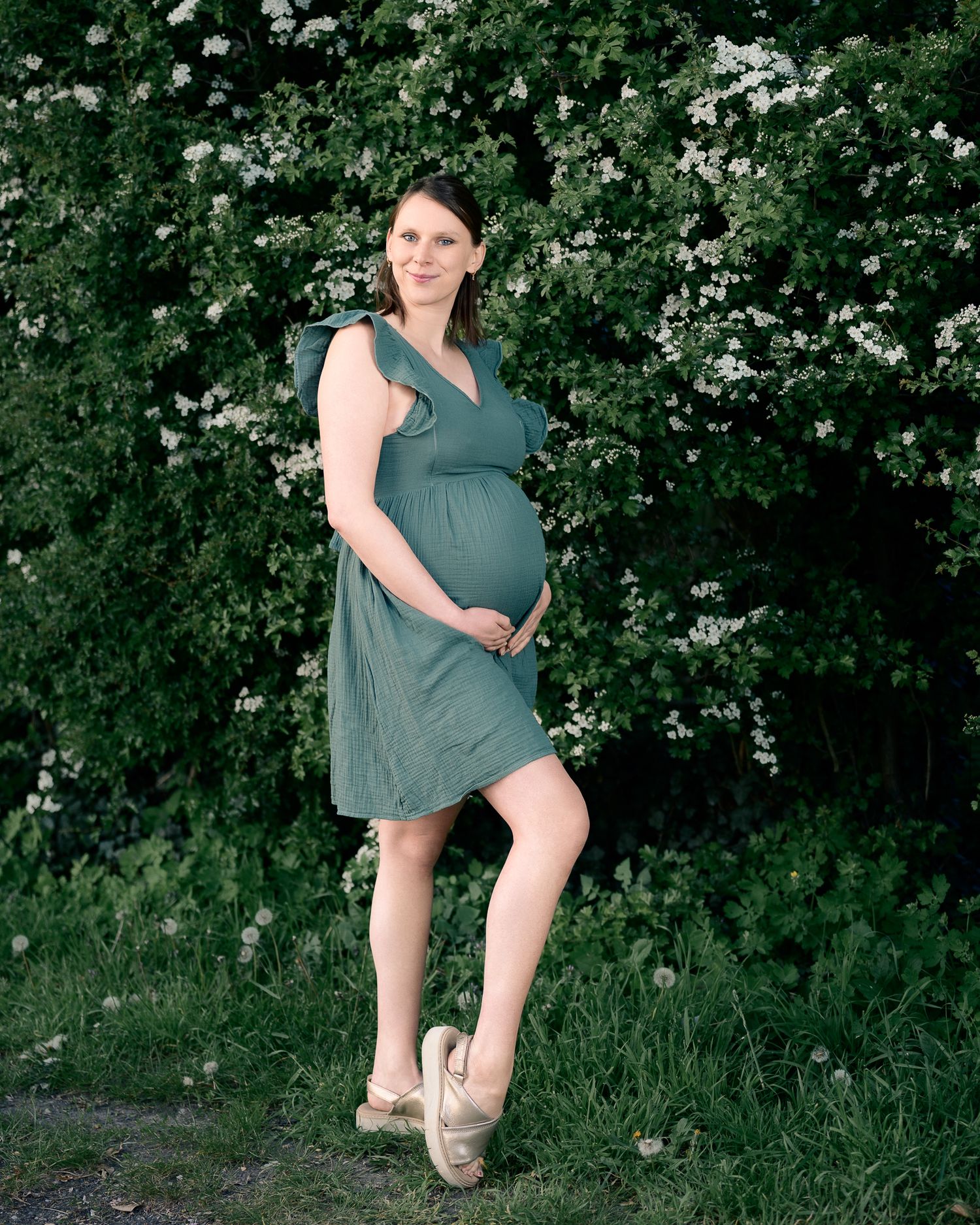 Pregnant woman in green dress posing outdoors with white flowering bushes, maternity photo session.