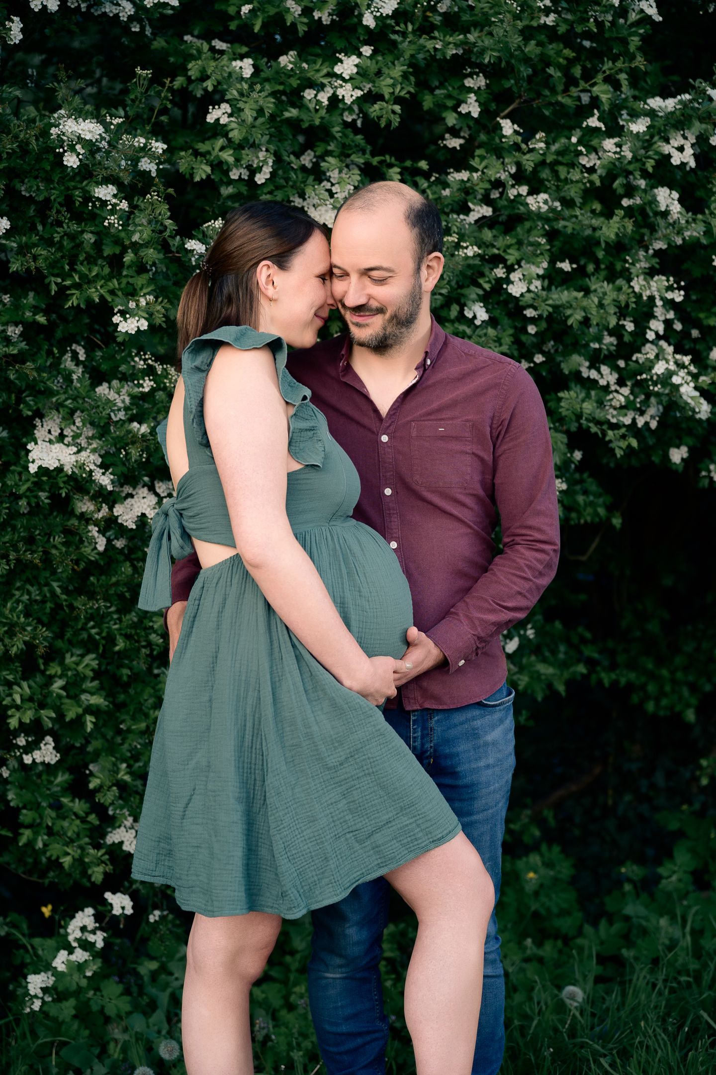Pregnant couple embracing outdoors near white flowering bushes, woman in green dress, man in burgundy shirt smiling.