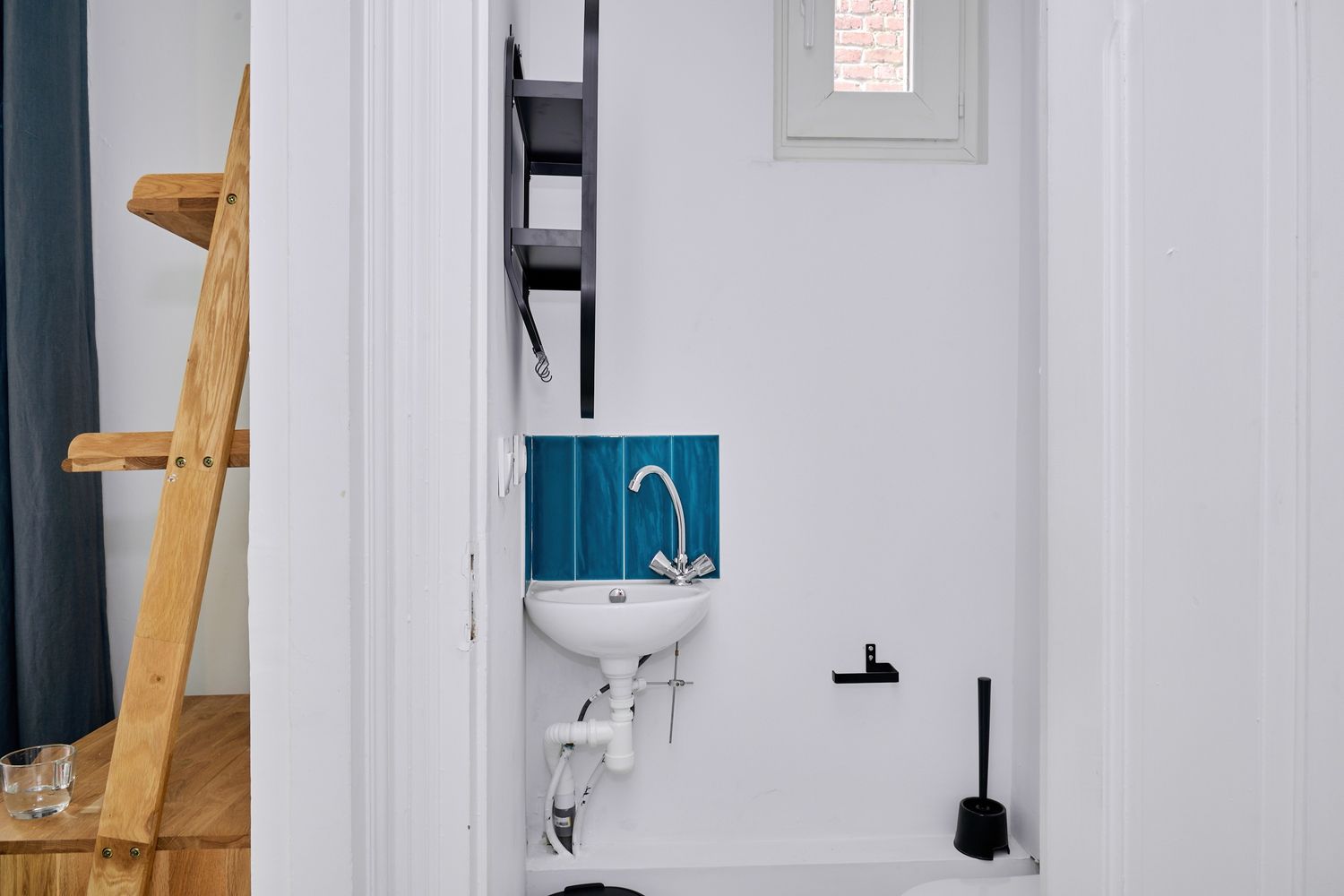 Compact white bathroom with blue tiled backsplash, wall-mounted sink, and wooden ladder shelf visible in doorway.