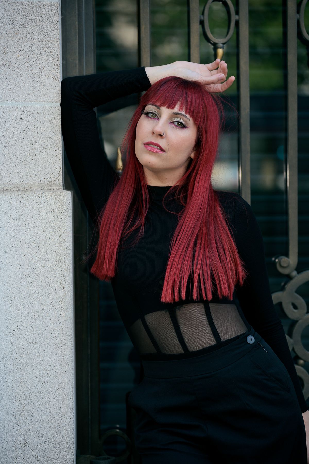 Red-haired woman in black sheer top leaning against ornate metal gate with hand raised.