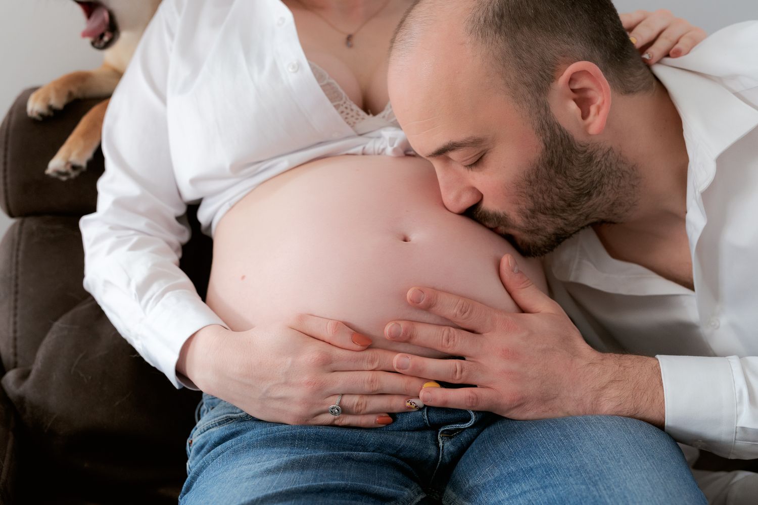 Expectant father kissing pregnant woman's bare belly while sitting together on a couch at home.