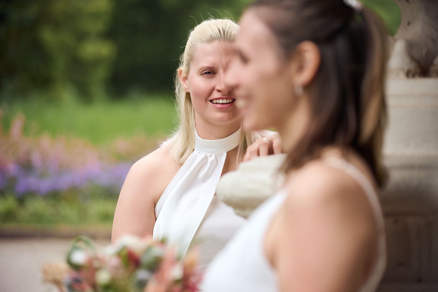 Two brides in white dresses smile at each other outdoors, with lush greenery and purple flowers in the background.