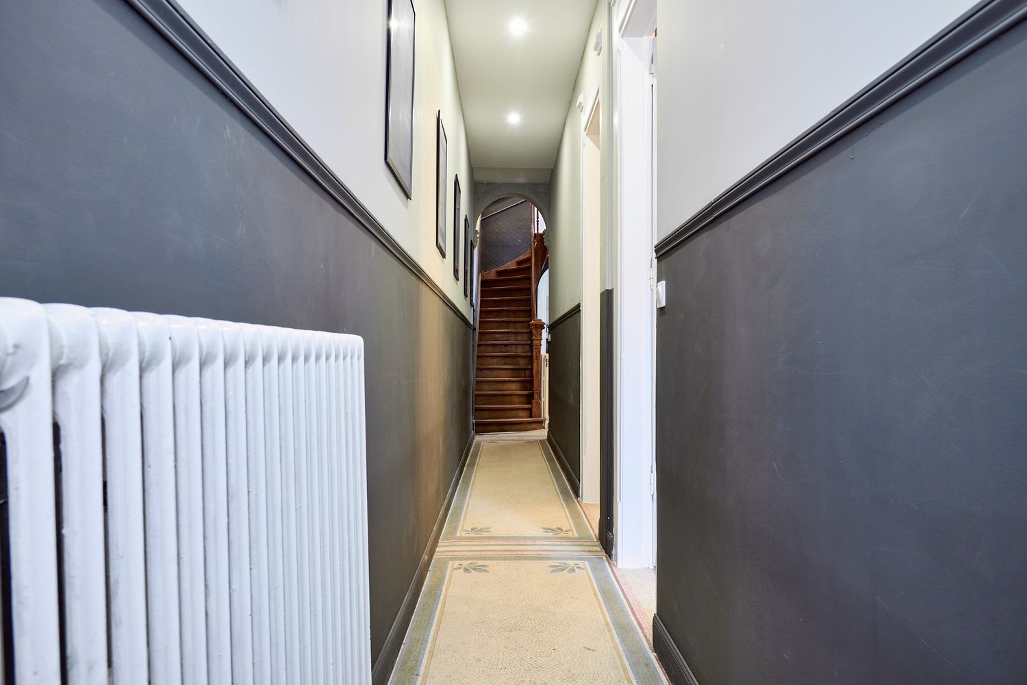 Narrow hallway with dark gray walls, white radiator, beige floor, and wooden staircase visible at the end.
