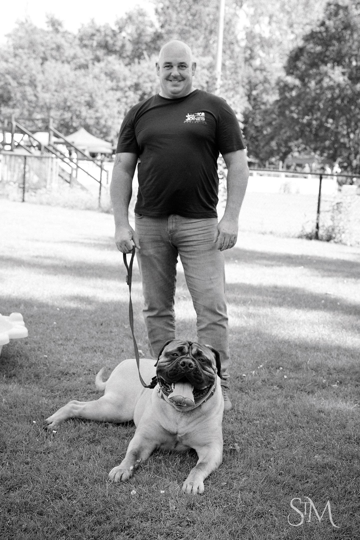 Bald man in black t-shirt smiling while standing with a pug on a leash in a grassy park.