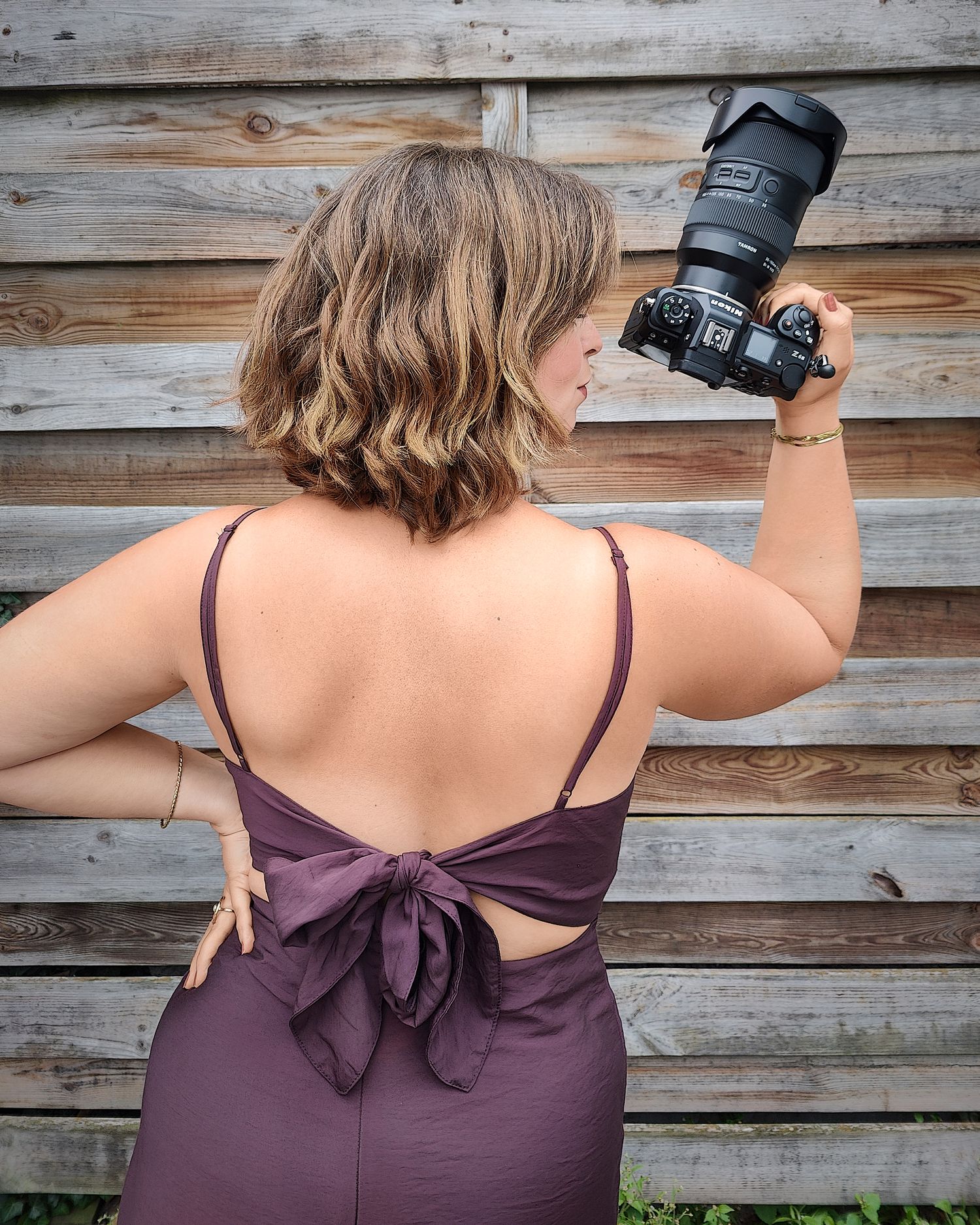 Woman in backless purple dress holding a DSLR camera up, standing in front of a rustic wooden fence.
