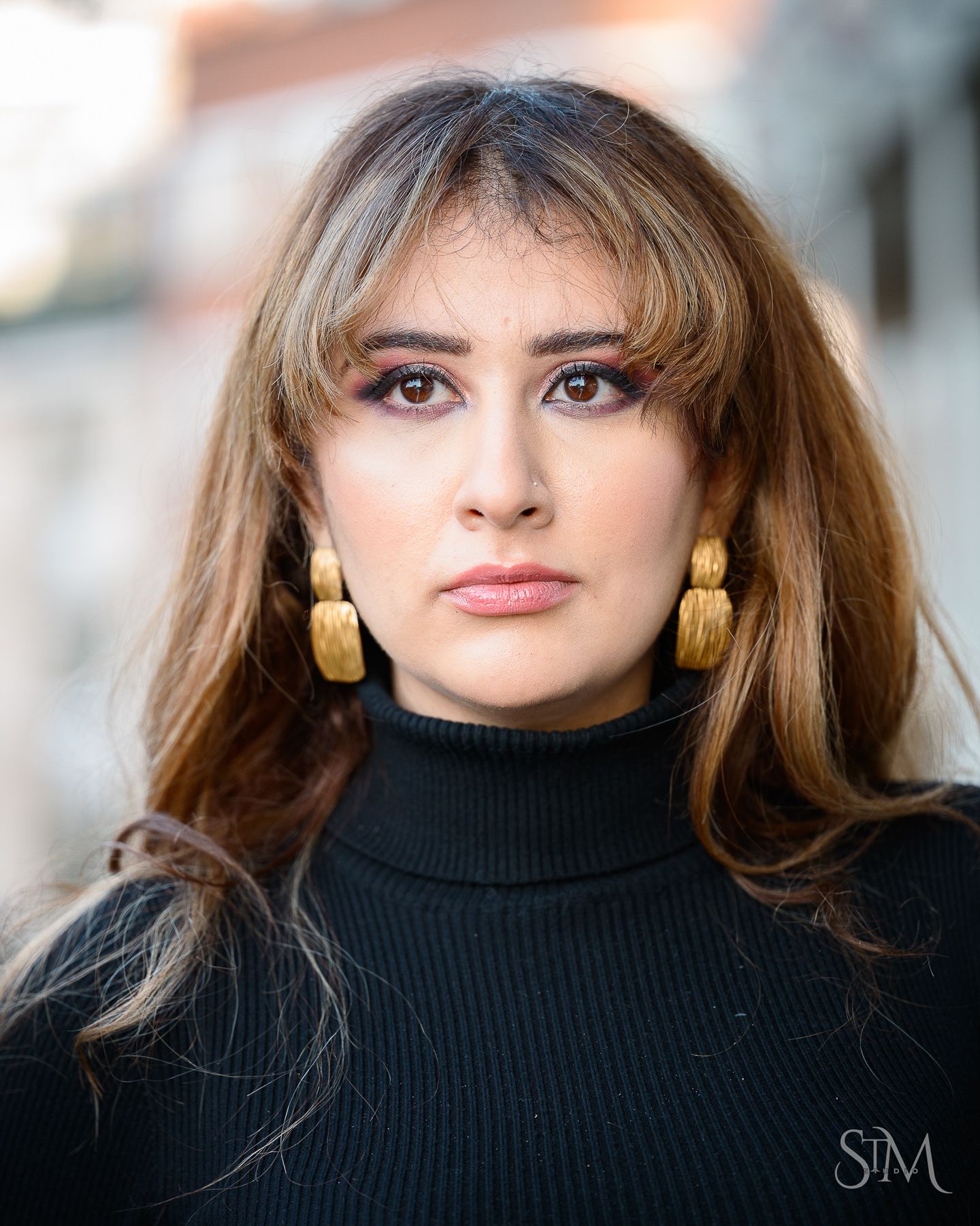 Young woman with bangs wearing gold earrings and black turtleneck sweater poses for outdoor portrait photo.
