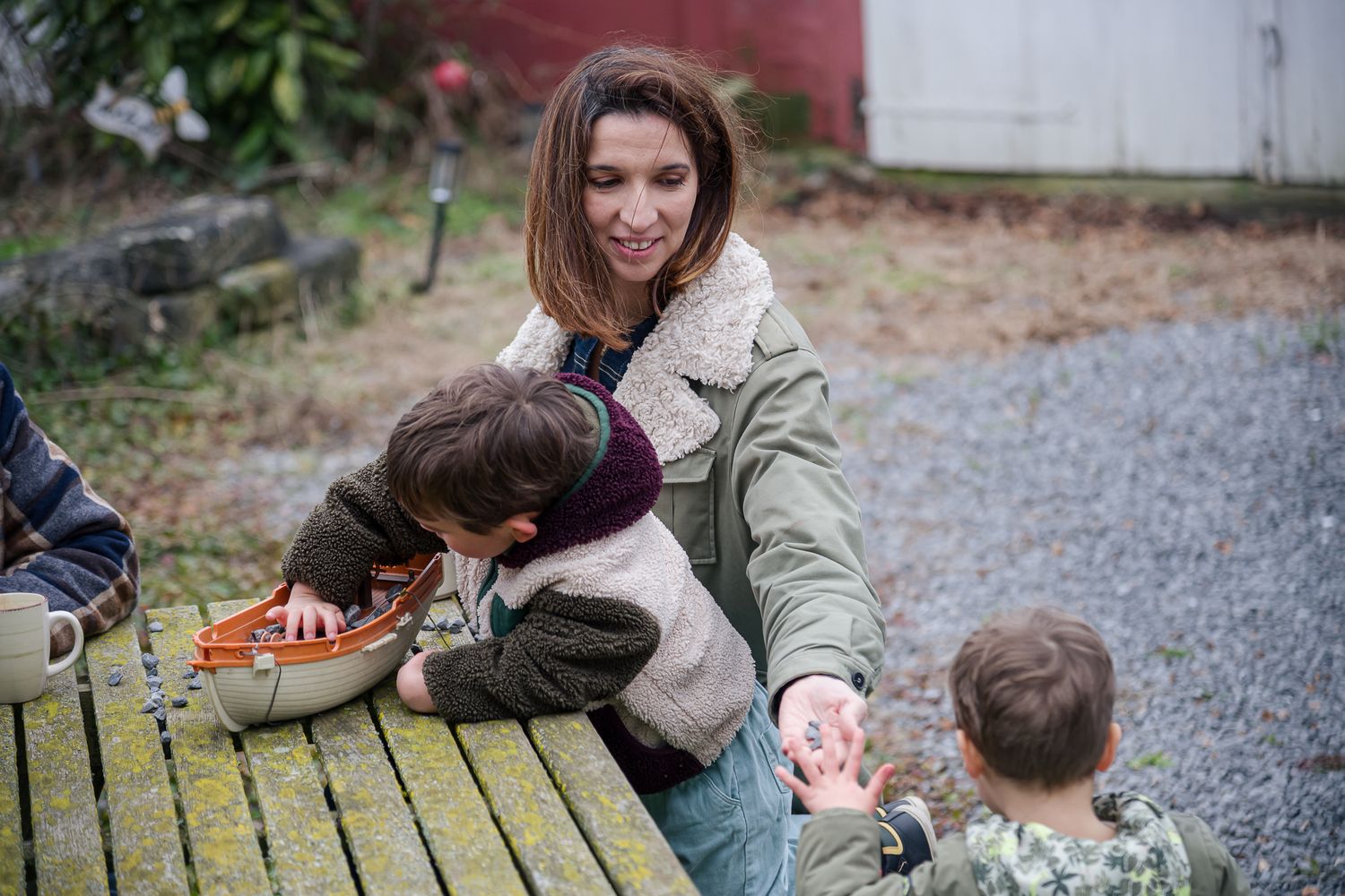 Mother helping two young children with a toy guitar on an outdoor bench.