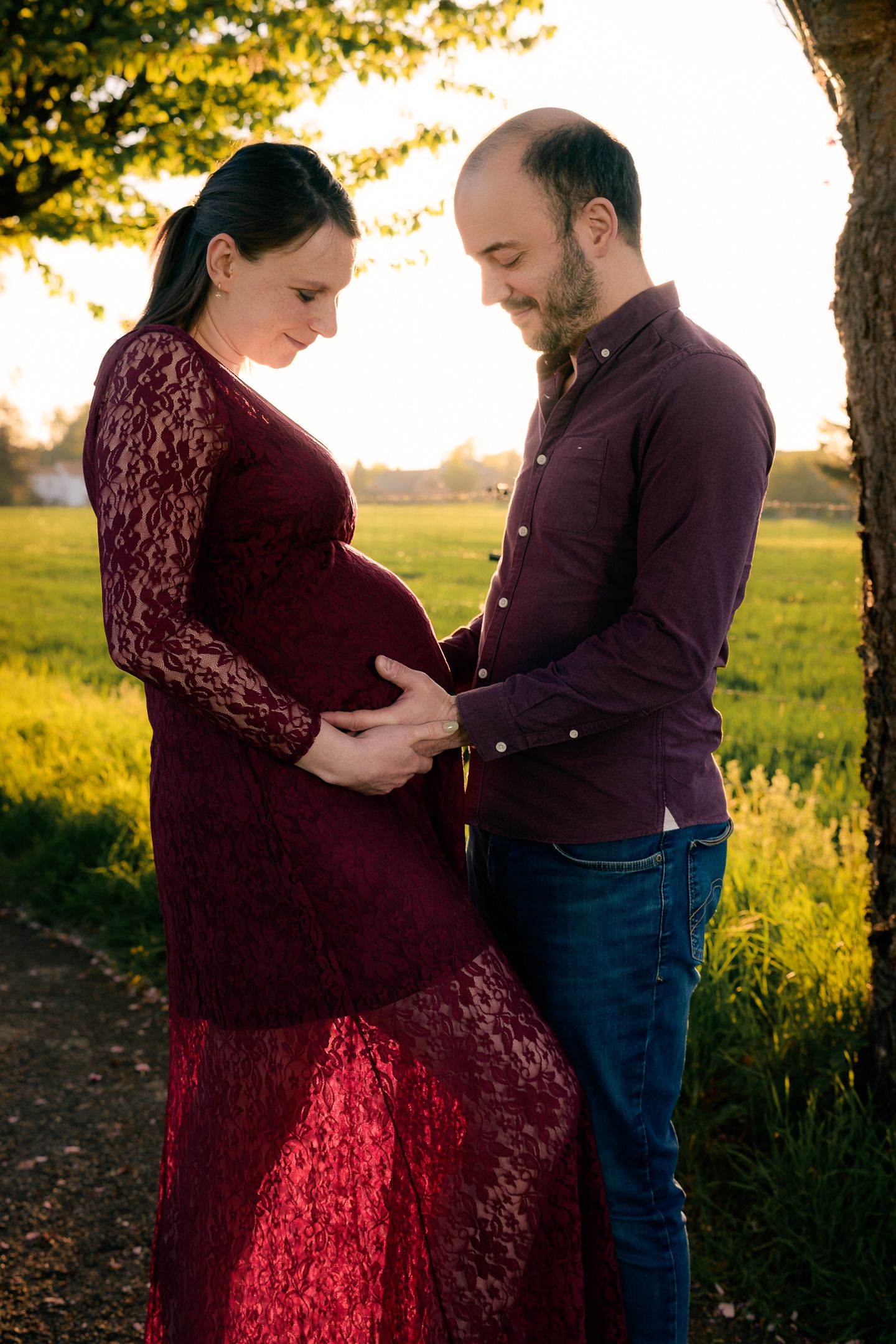 Couple tenderly cradles pregnant belly outdoors at golden hour, woman in red lace dress, man in burgundy shirt and jeans.