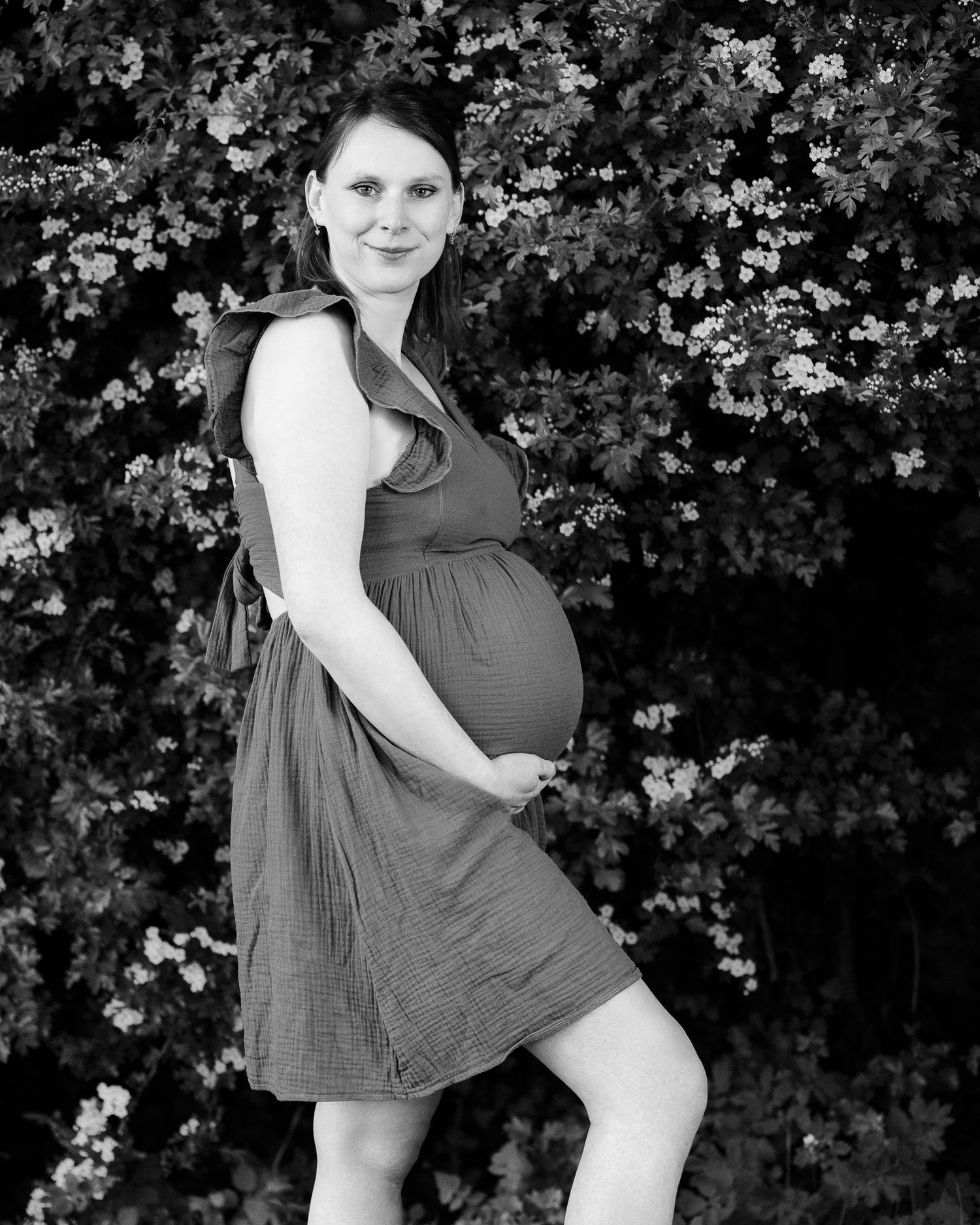Black and white photo of a pregnant woman in a dress cradling her baby bump, posing outdoors near flowering bushes.