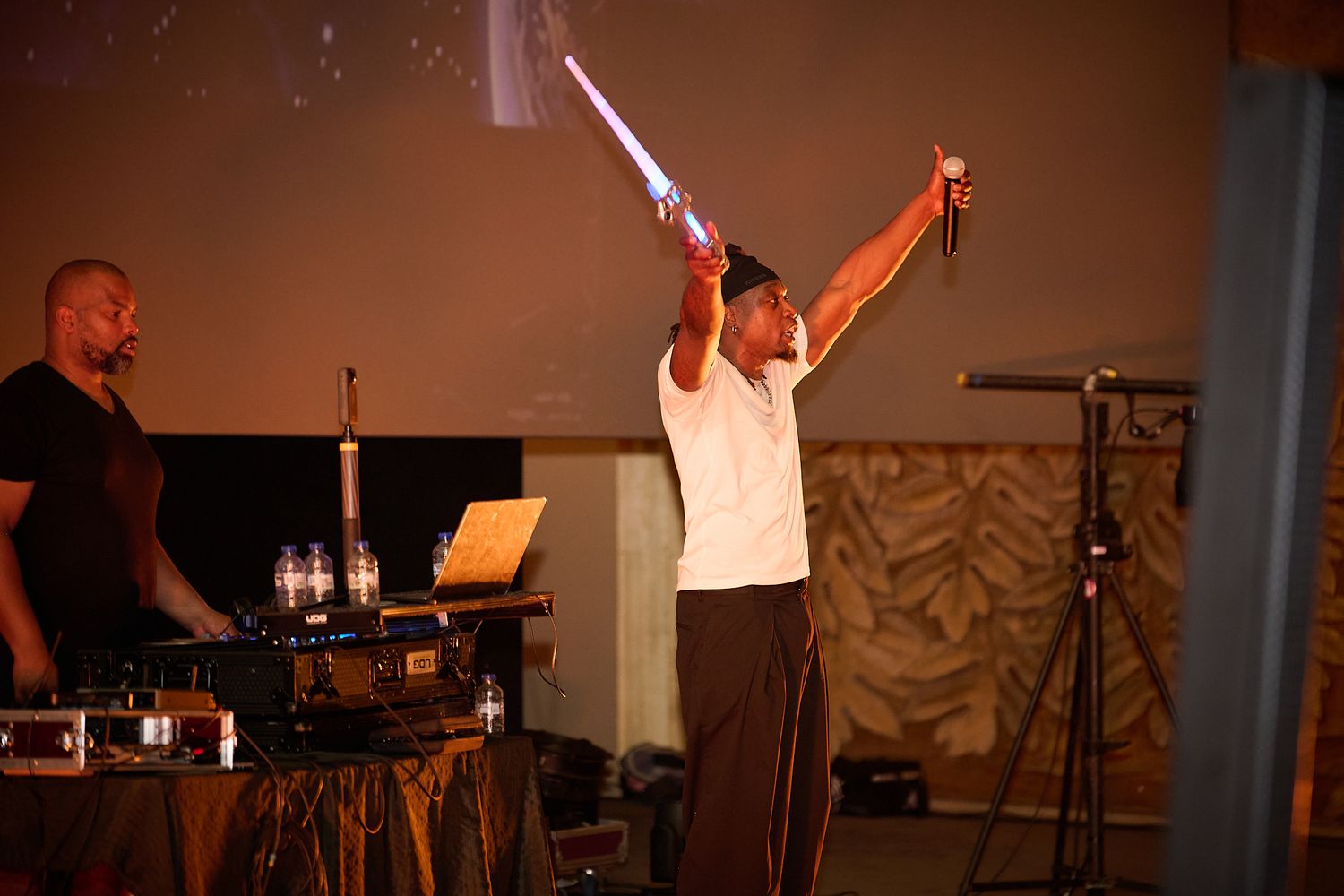 Performer raises glowing lightsaber on stage while DJ operates equipment at an indoor event.