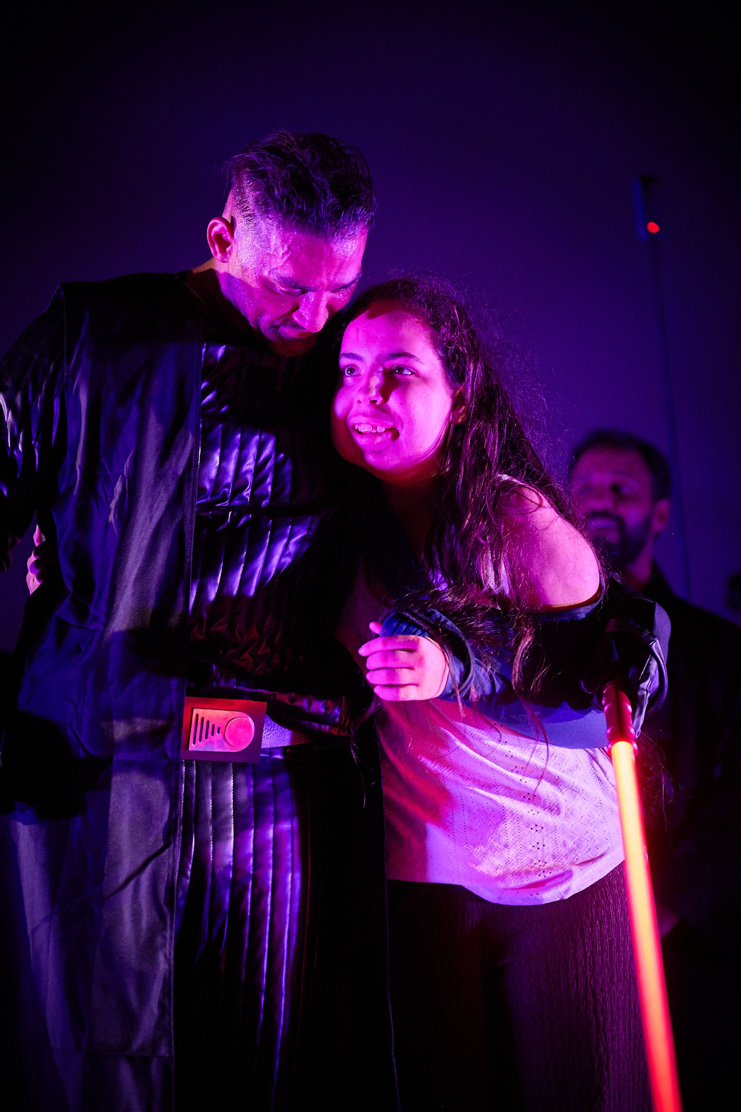 Two performers embrace on stage under purple and red lighting during a dramatic live show.