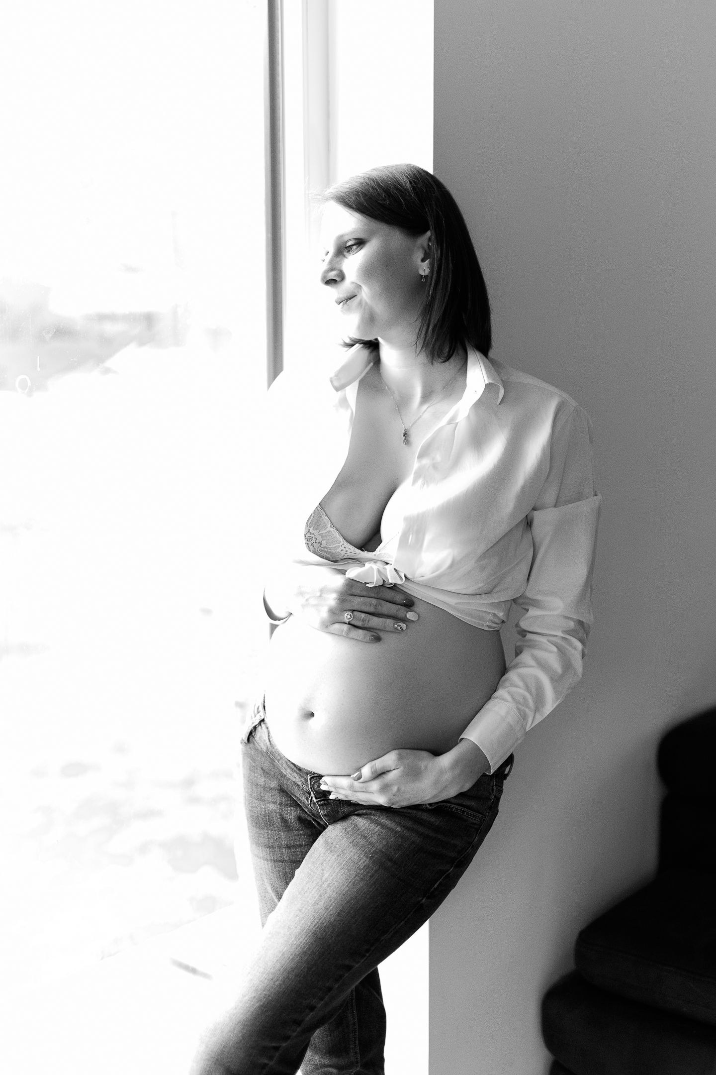 Black and white photo of a smiling pregnant woman in open white shirt and jeans, cradling her baby bump by a window.