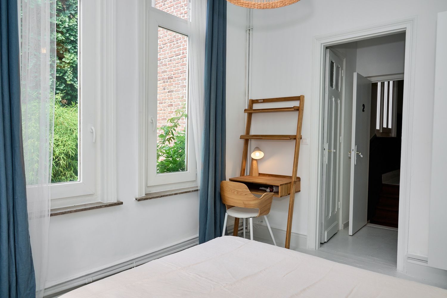 Bright bedroom with blue curtains, wooden desk, stool, and open door revealing a hallway in a modern apartment.