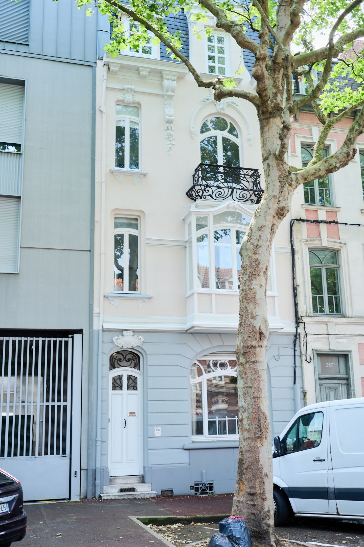 Elegant white French townhouse with ornate balcony, arched windows, and decorative ironwork, flanked by a large plane tree.