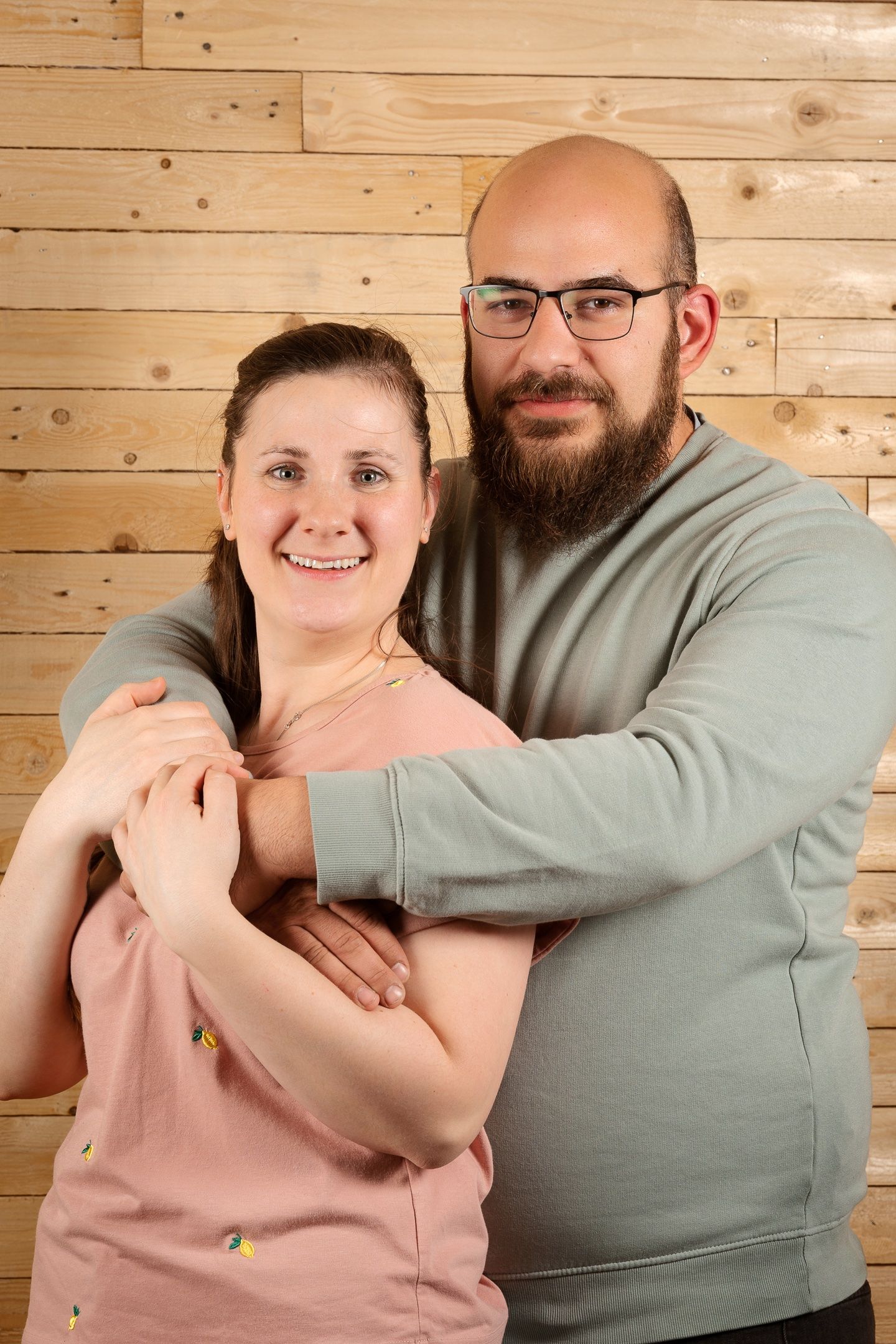 Smiling couple embracing against wooden wall, bearded man with glasses hugging woman in pink top.