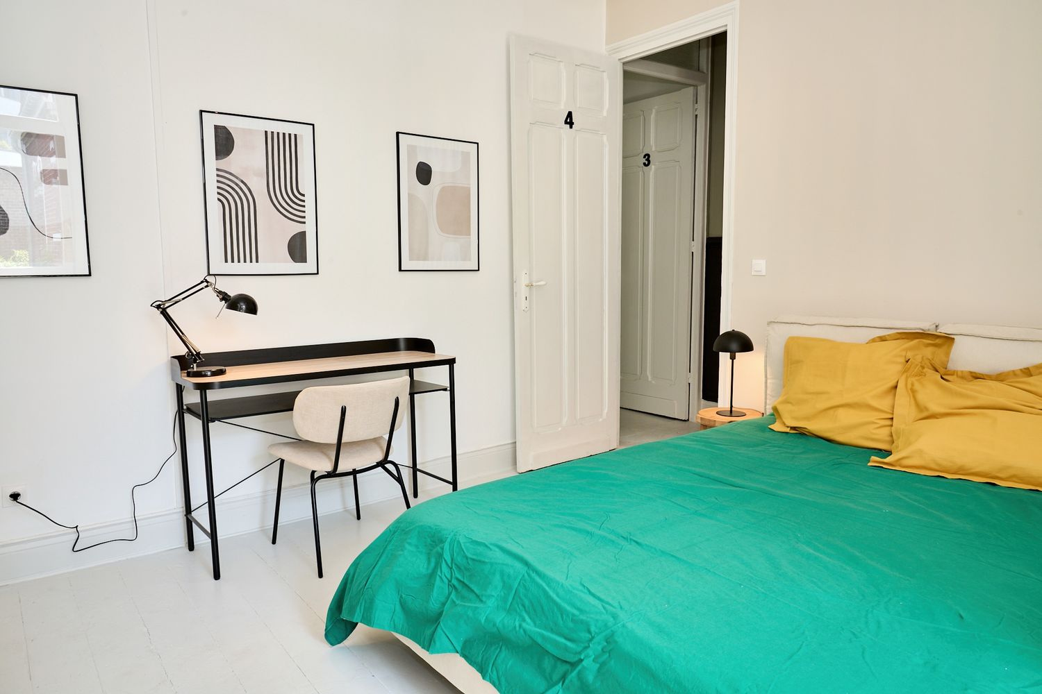 Modern bedroom with green bedspread, yellow pillows, minimalist desk, and abstract wall art in a bright white room.