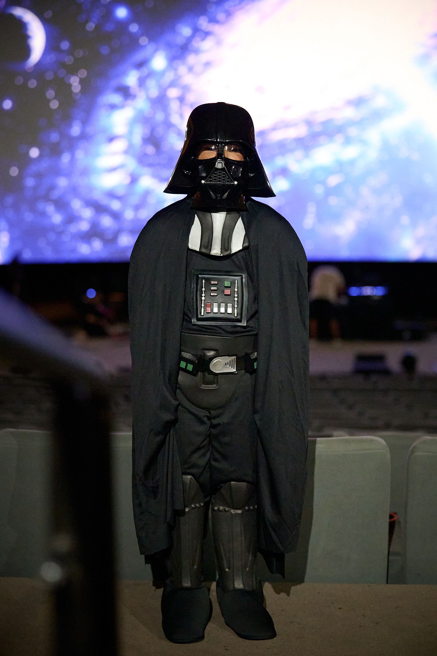Child dressed in Darth Vader costume stands in front of a Star Wars themed backdrop at a cinema event.