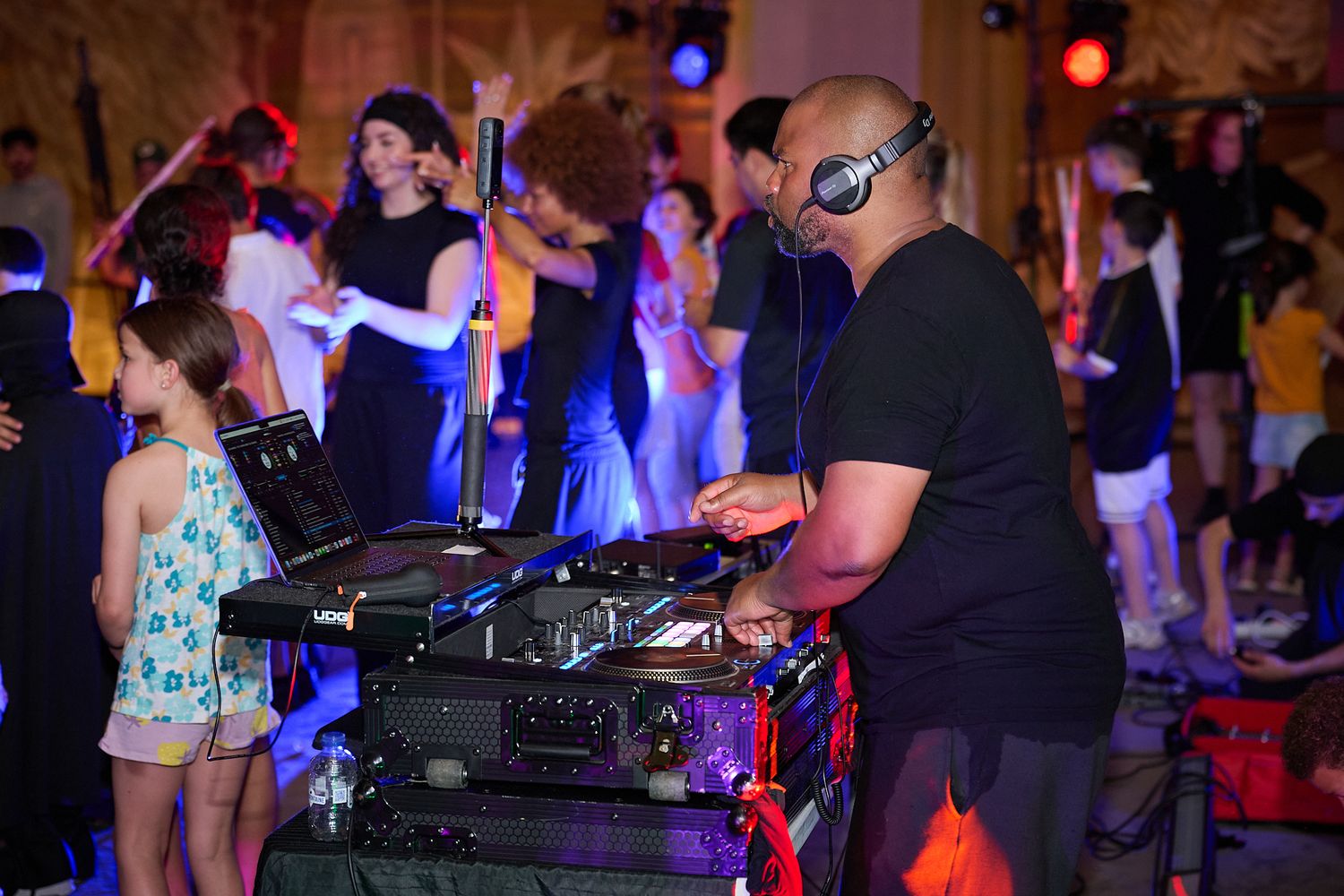 A DJ wearing headphones operates turntables at a lively party with dancers in the background.
