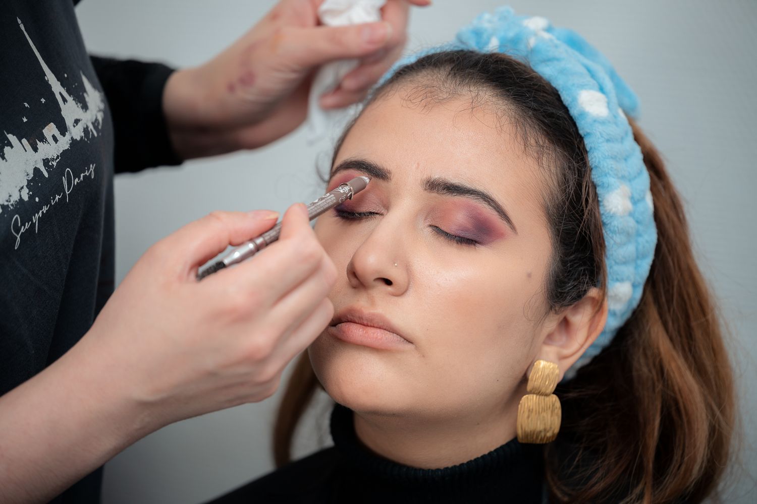 Professional makeup artist applying eyeshadow to a young woman wearing a blue headband and gold earrings.