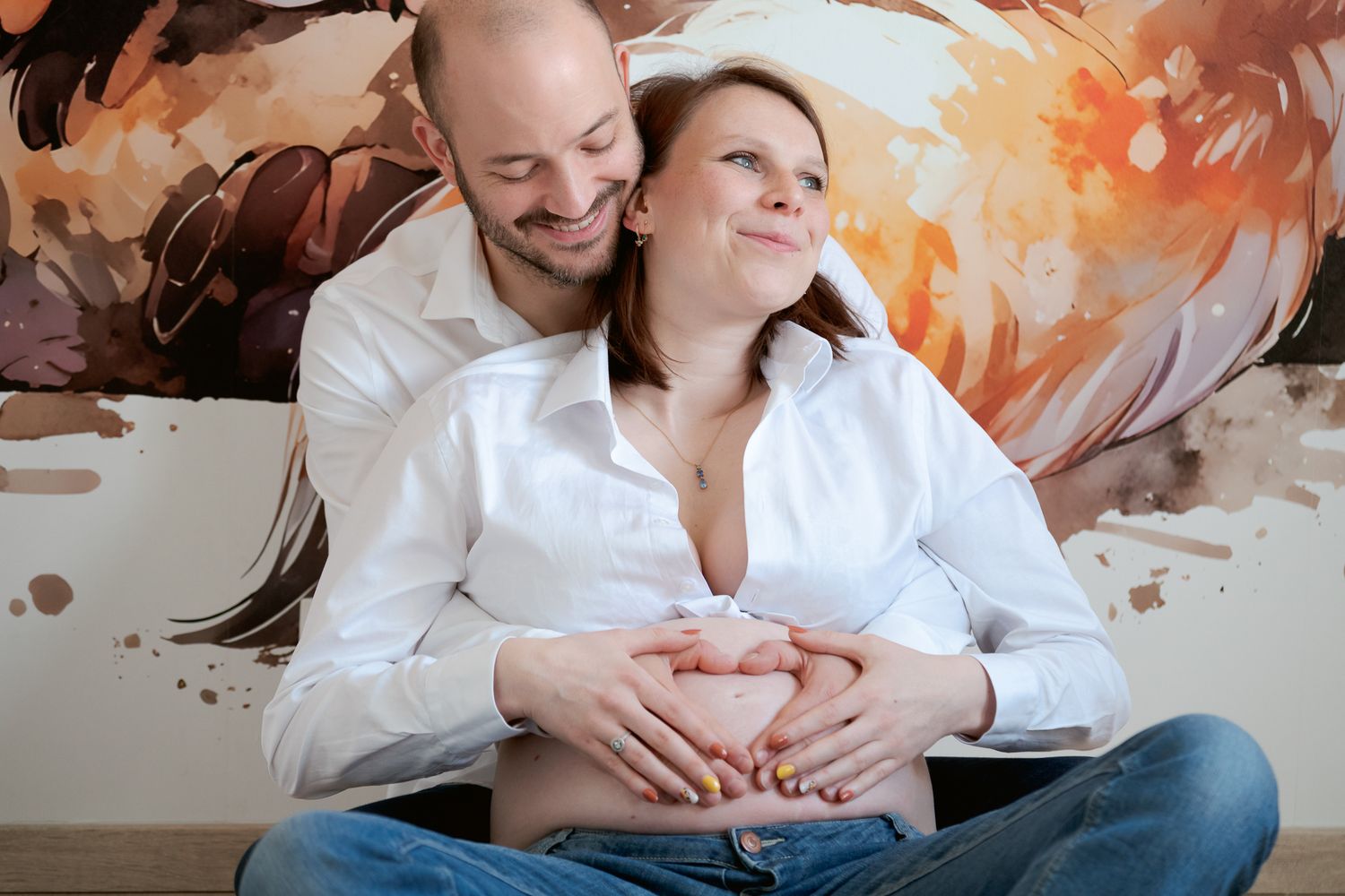 Happy couple expecting a baby, man embracing pregnant woman forming heart shape on belly, sitting together smiling.