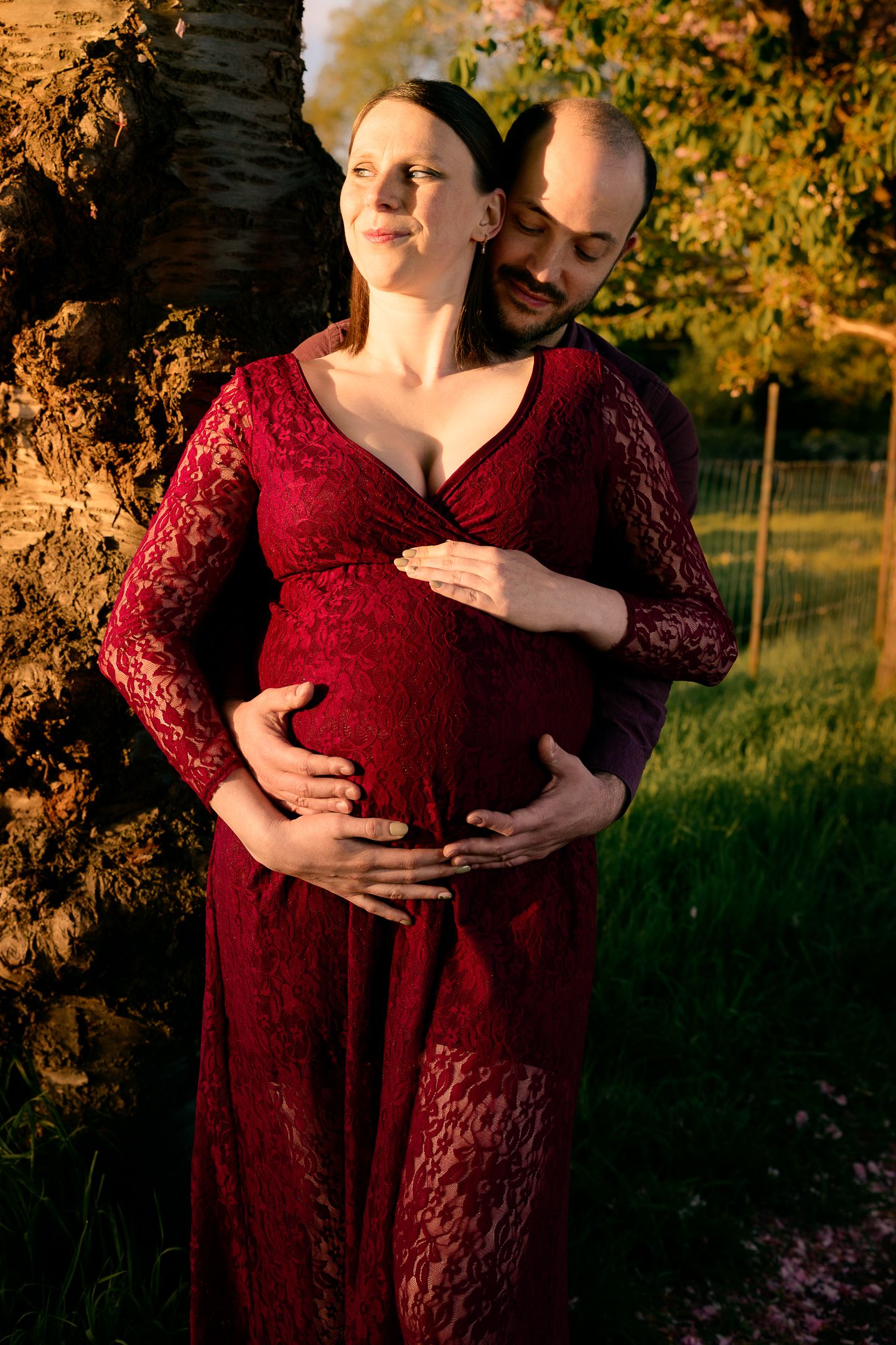 Couple embracing outdoors at golden hour, pregnant woman in red lace dress leaning against tree in garden.