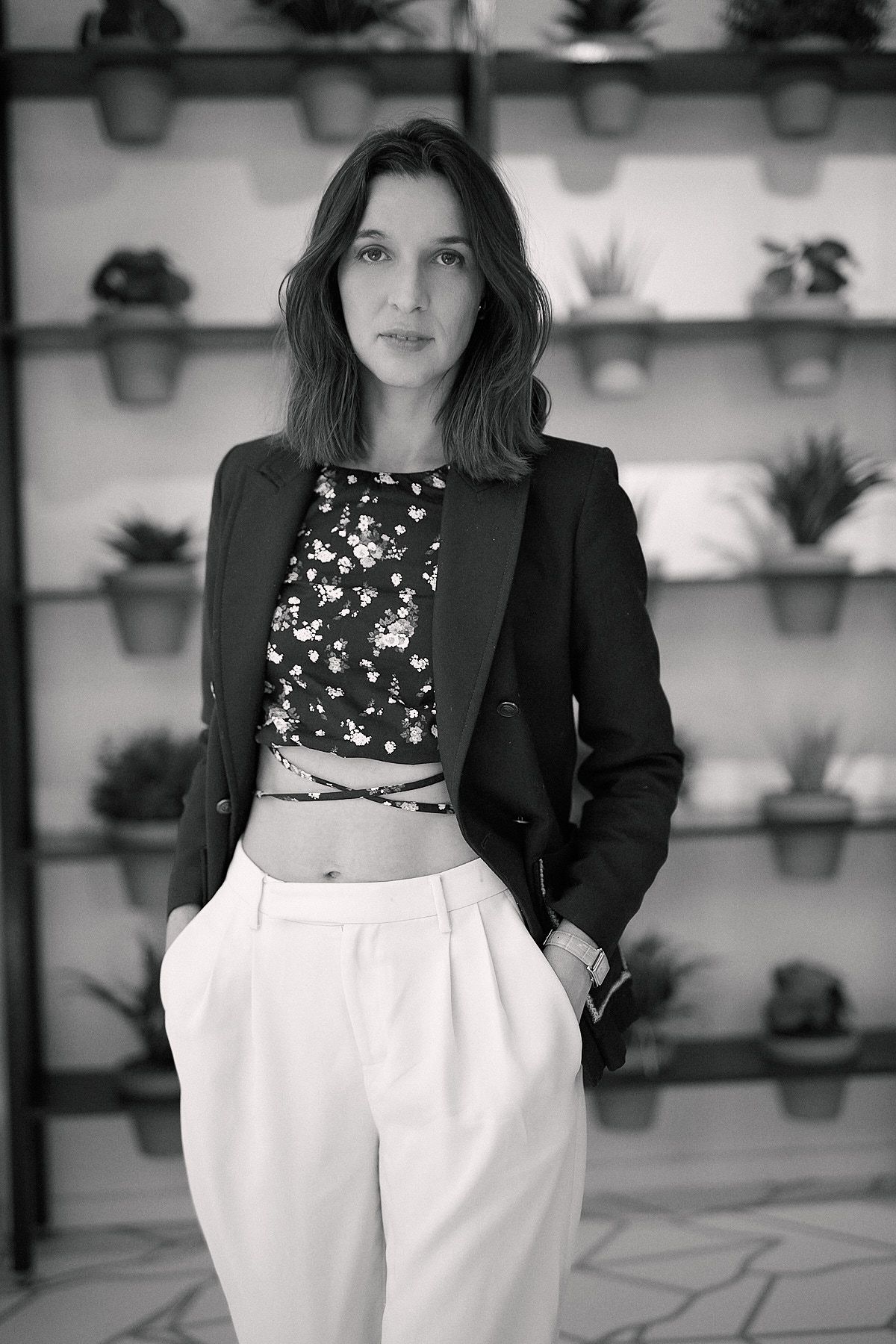 Black and white portrait of a young woman in blazer and wide-leg trousers, posing before shelves of potted plants.