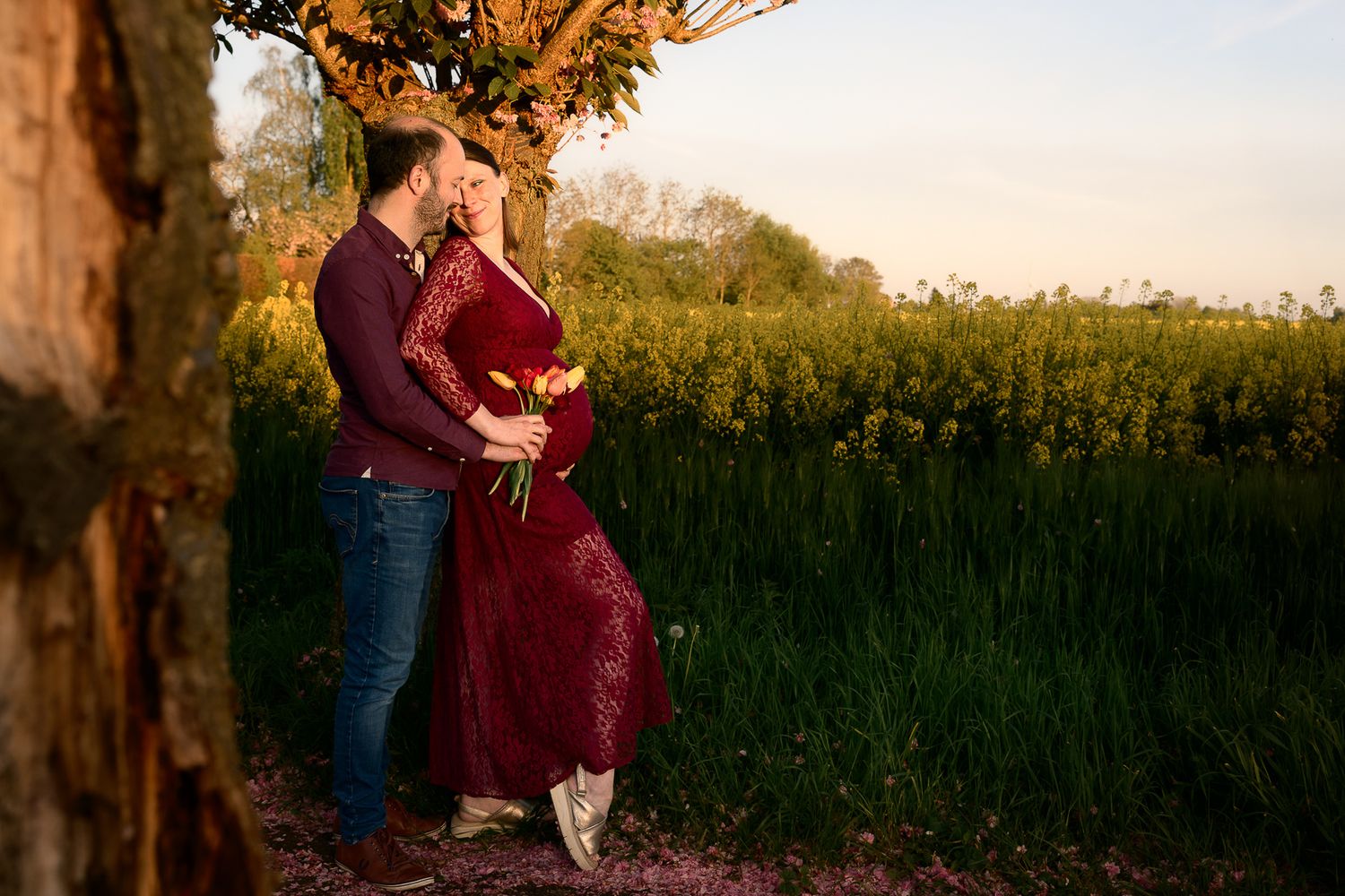 Couple embracing near a tree in a golden field, woman in red lace maternity dress, romantic outdoor pregnancy photo.