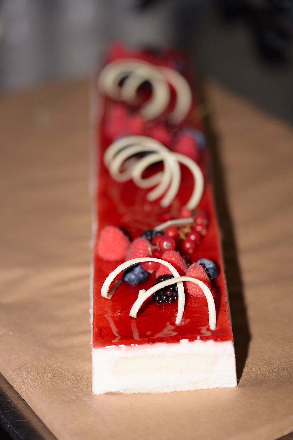 Elegant rectangular berry dessert with red glaze, fresh raspberries, blueberries, and white chocolate spiral decorations.