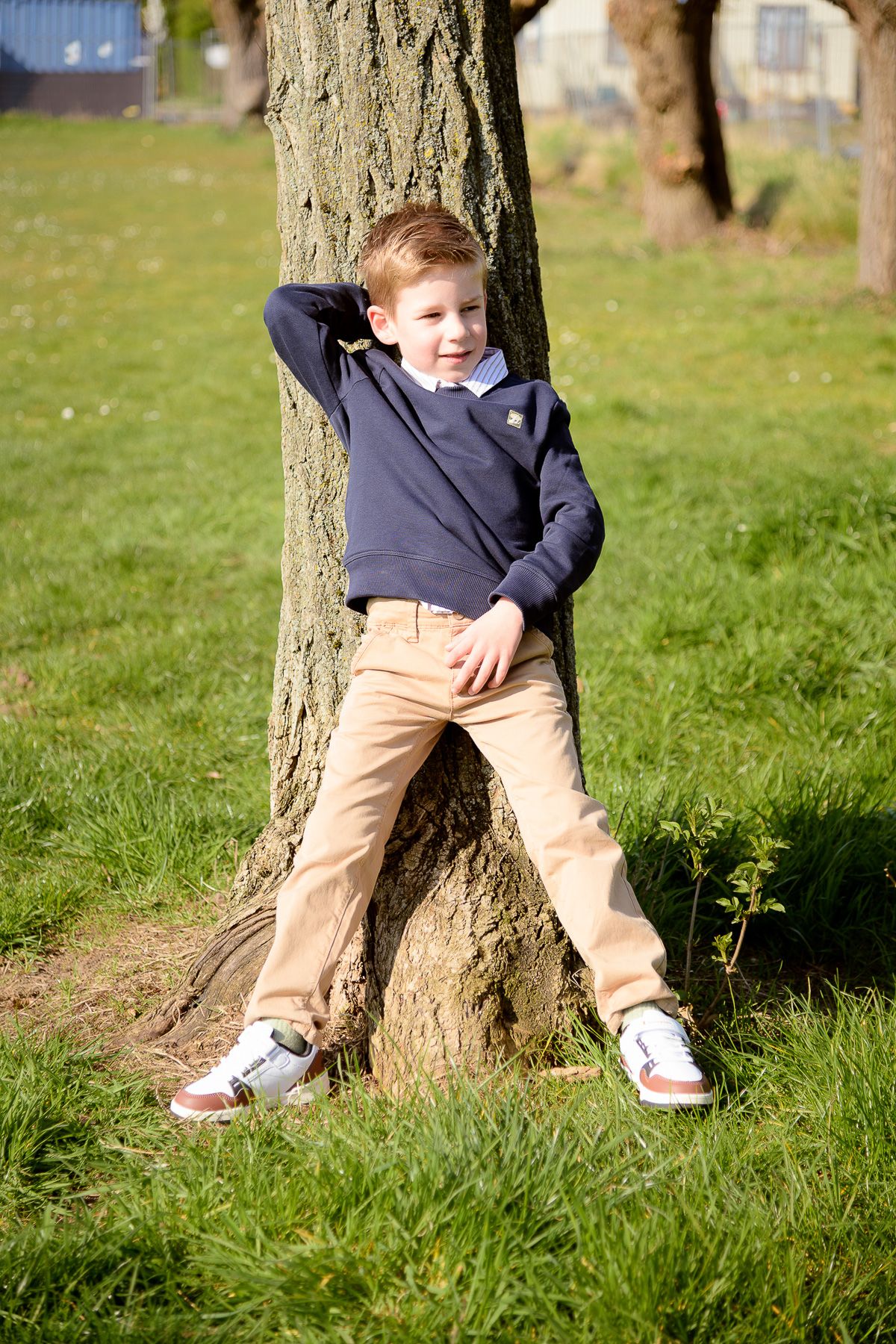 A young boy in a navy sweater and khaki pants leans against a tree trunk in a sunny green park.