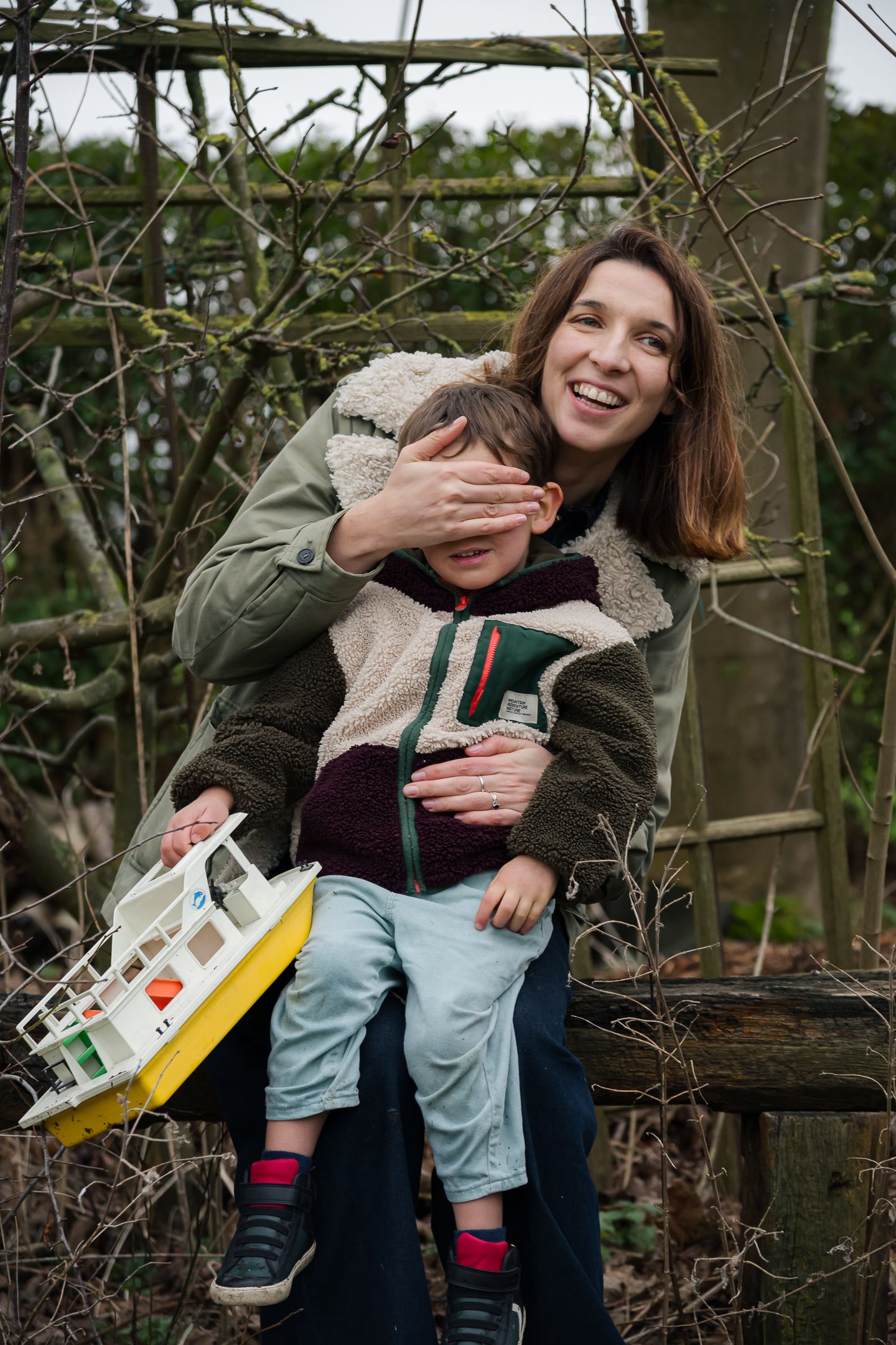 Smiling mother playfully covering her young childs eyes while sitting outdoors on a bench.
