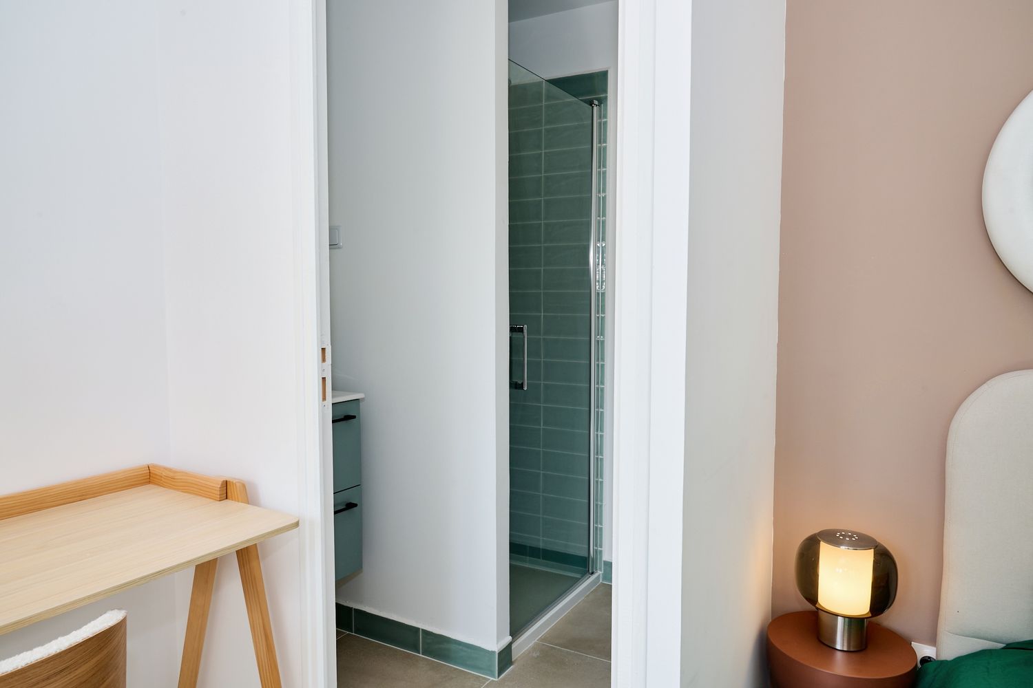 Modern hotel room with open bathroom door revealing green tiled shower, wooden desk, and stylish bedside lamp.