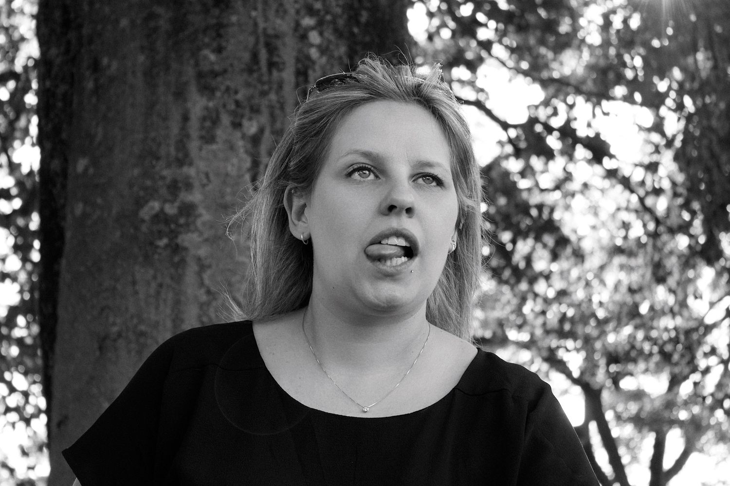 Black and white portrait of a young blonde woman outdoors near a tree, looking upward with mouth open.