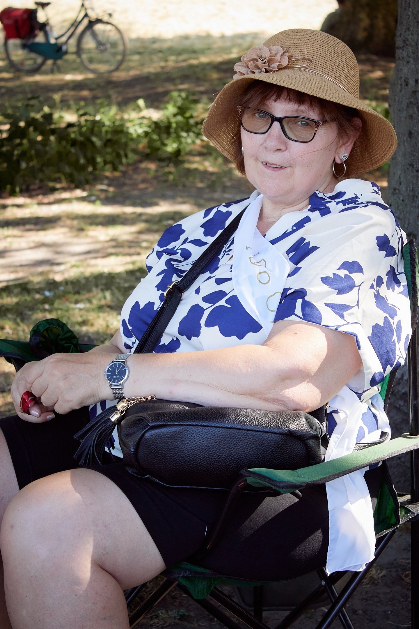 Elderly woman in straw hat, glasses, and floral blouse sitting in folding chair outdoors in shaded park area.