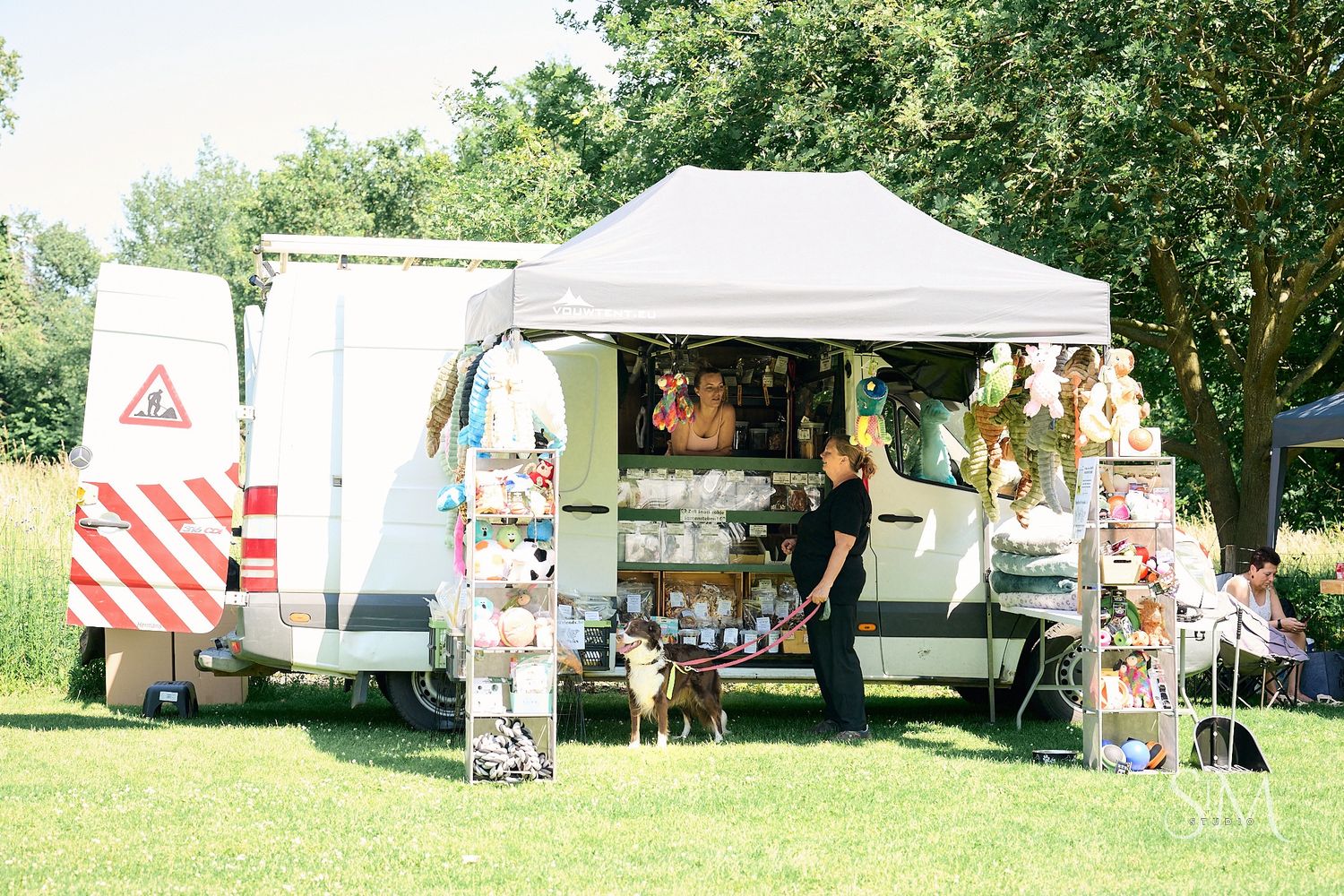 Outdoor market stall with white van and canopy tent displaying colorful goods on a sunny green field.