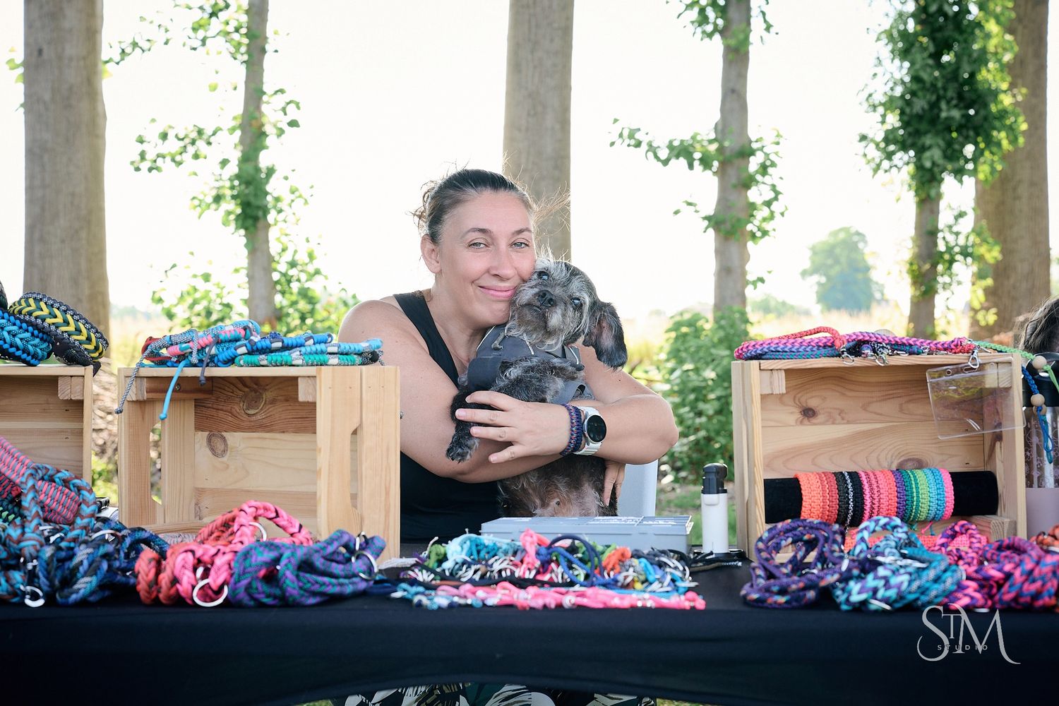 Woman smiling at outdoor market stall holding a rabbit surrounded by colorful pet leashes and accessories on display table.