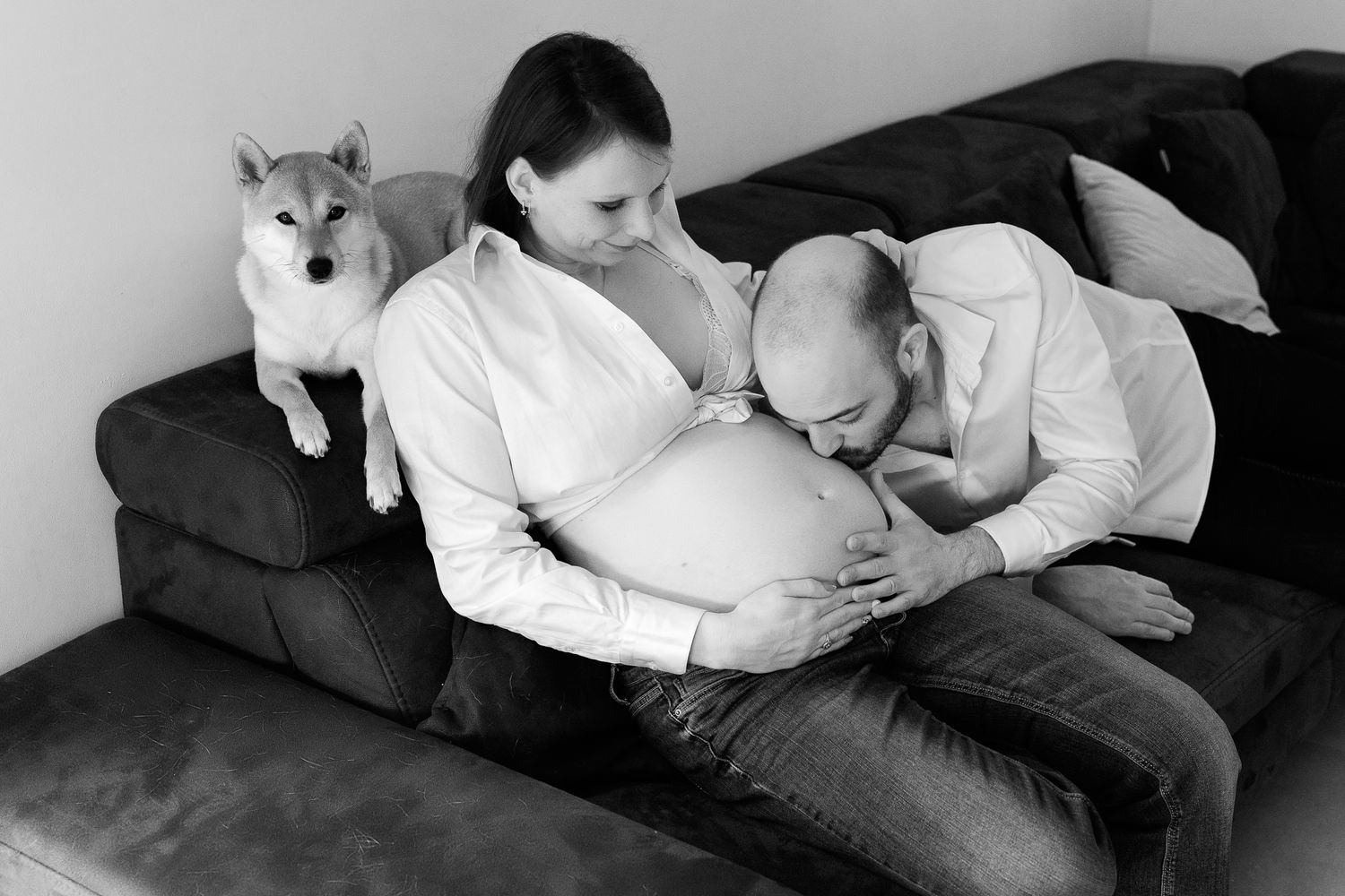 Black and white photo of man kissing pregnant woman's belly on sofa with dog watching.