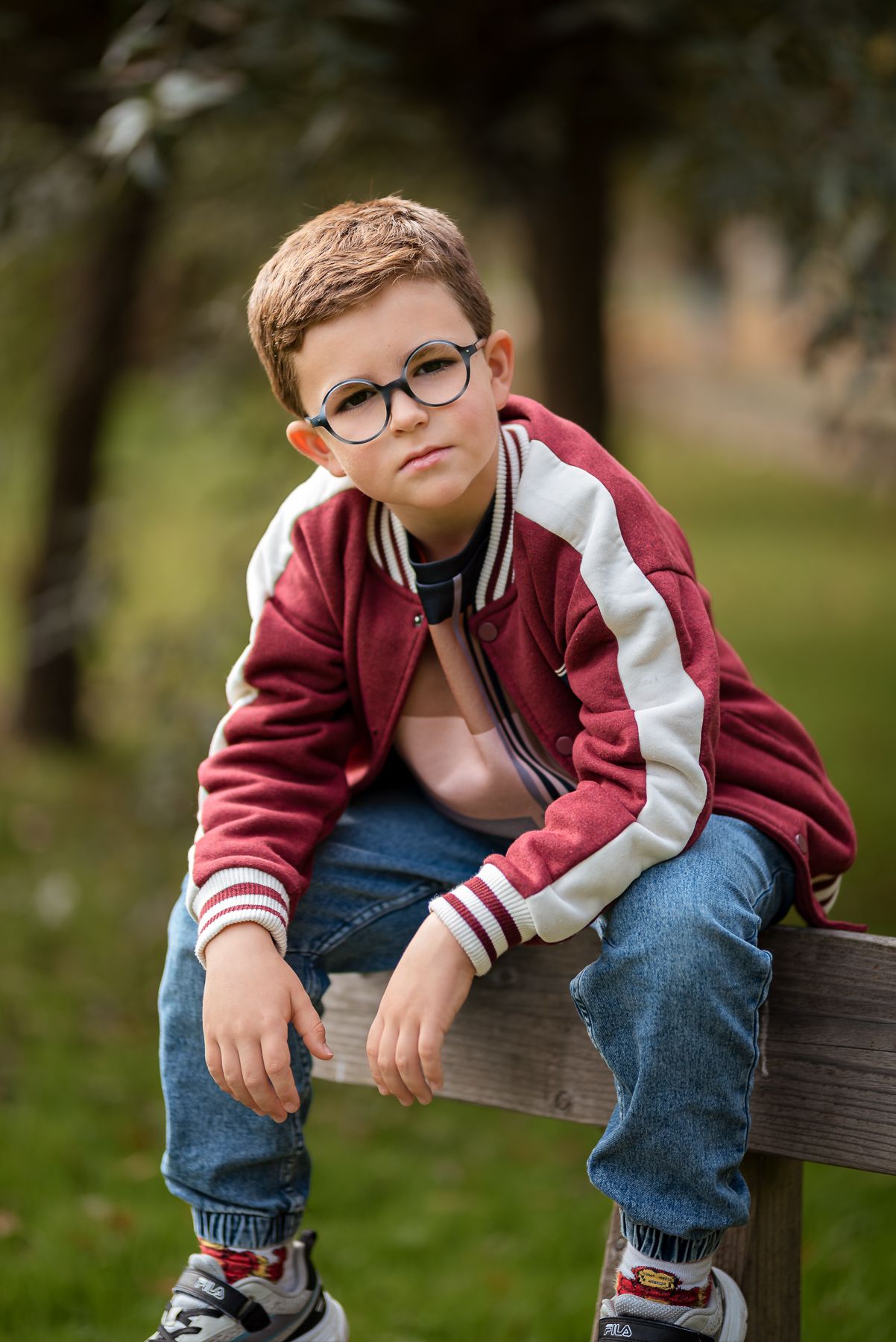 A young boy in a red varsity jacket and glasses sits on a wooden bench in a park.