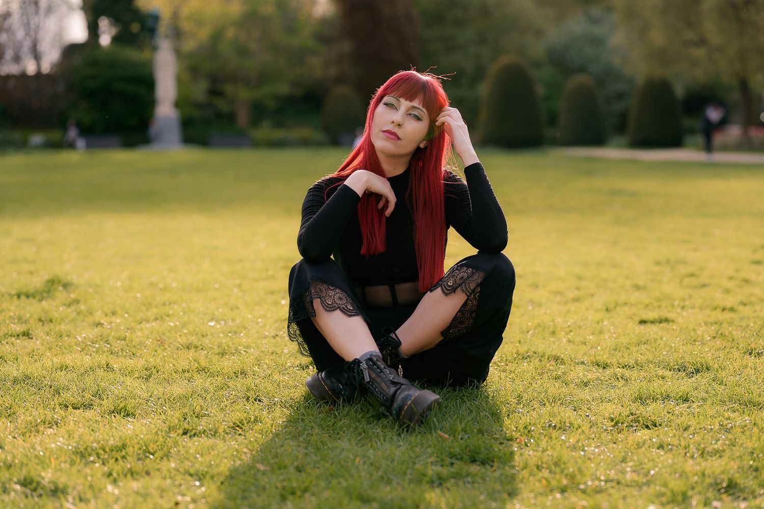 Young woman with red hair in black outfit sitting cross-legged on green grass in a park during golden hour.