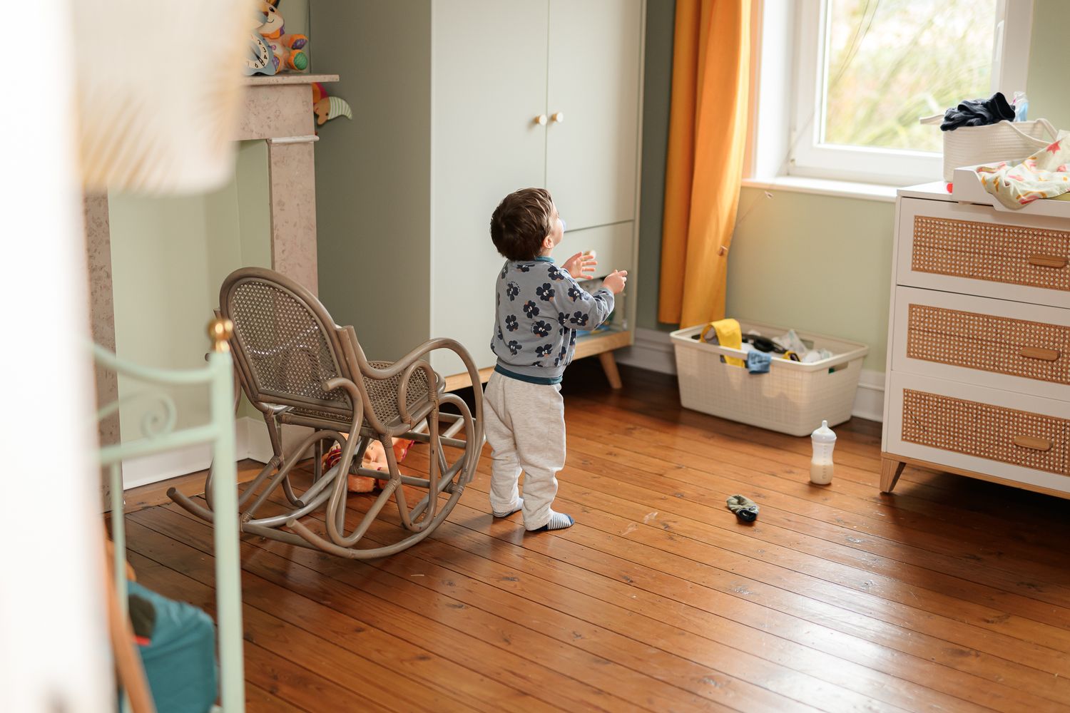 Young toddler playing alone in a nursery room with a rocking chair and wooden dresser.