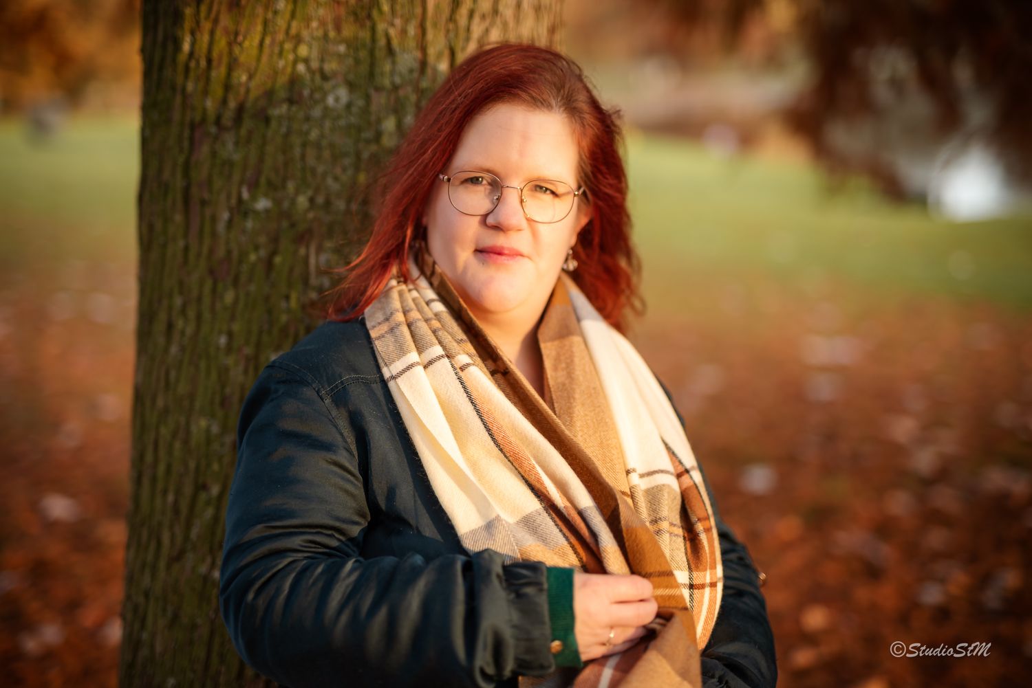 Red-haired woman with glasses holding green cup leaning against tree in golden autumn park light.